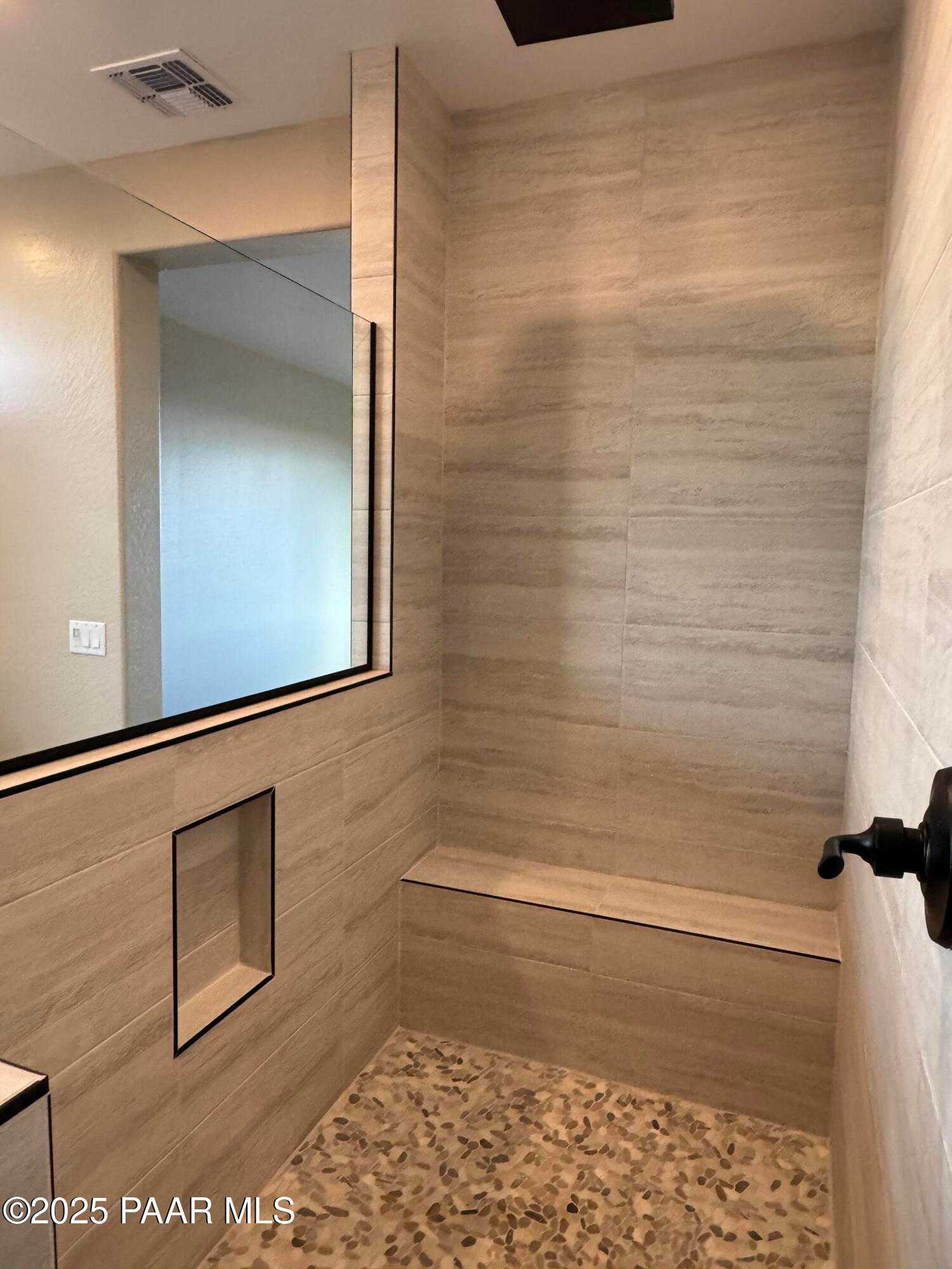 Luxurious walk-in shower with beige tile walls, frameless glass enclosure, bench seat, and pebble floor in Evermore Homes The Sunrise A, Prescott Valley, AZ