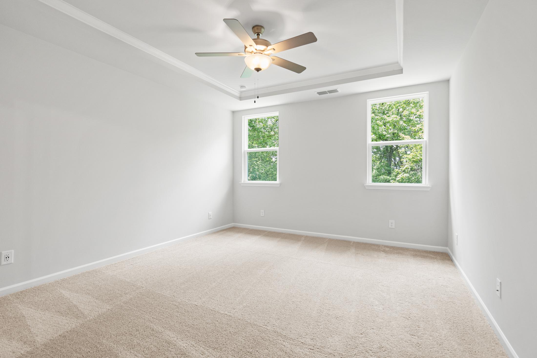 Spacious empty bedroom in The Ash B home at Wehunt Meadows with beige carpet, ceiling fan, tray ceiling, and large windows overlooking greenery