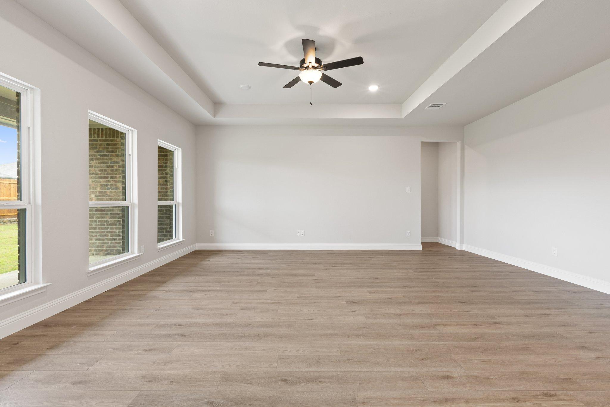 Spacious living room in The Harrison home with tray ceiling, ceiling fan, hardwood floors, and large windows
