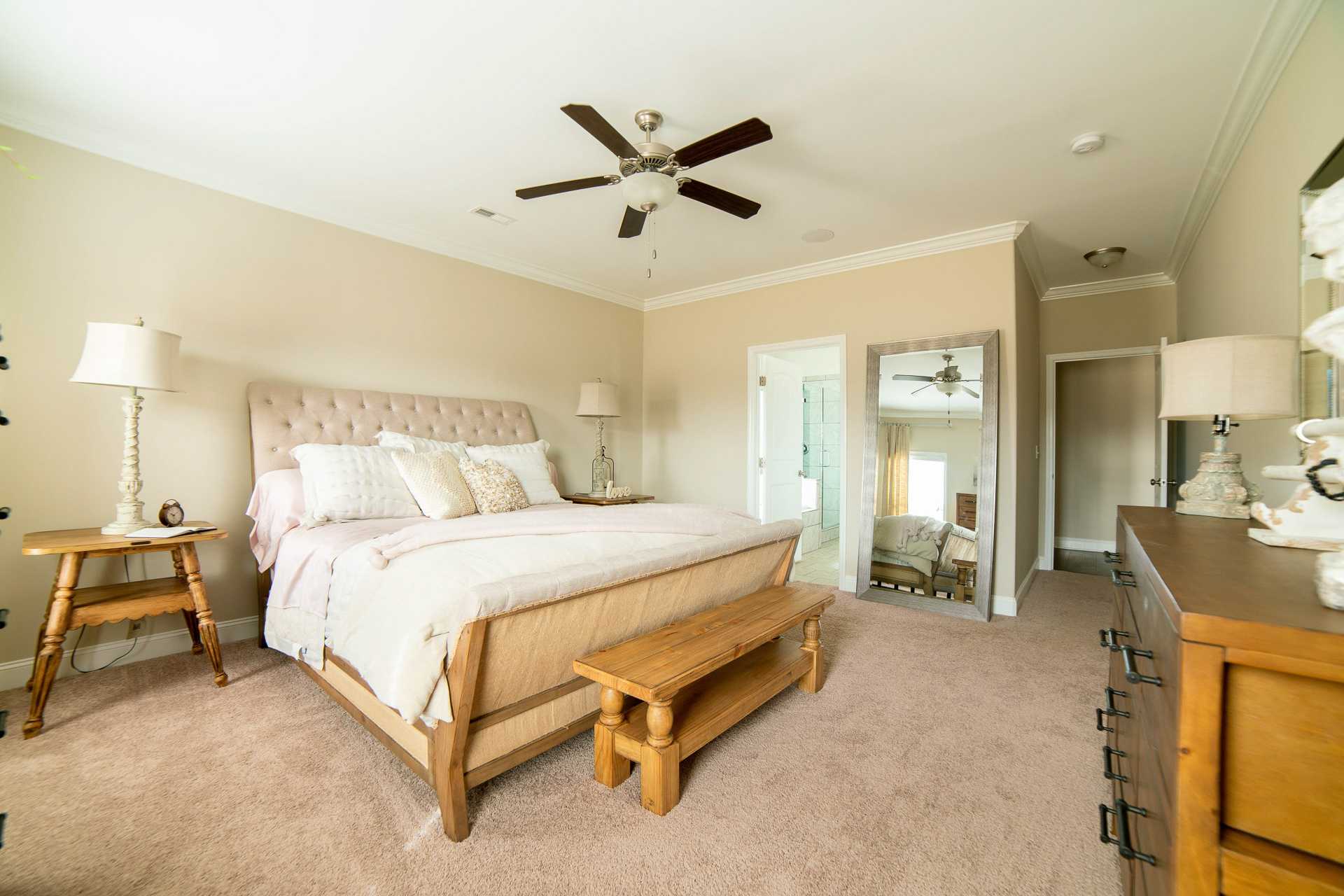 Spacious master bedroom at The Dairy at Oak Grove in South Huntsville AL with beige walls, upholstered bed, ceiling fan, and wooden bench