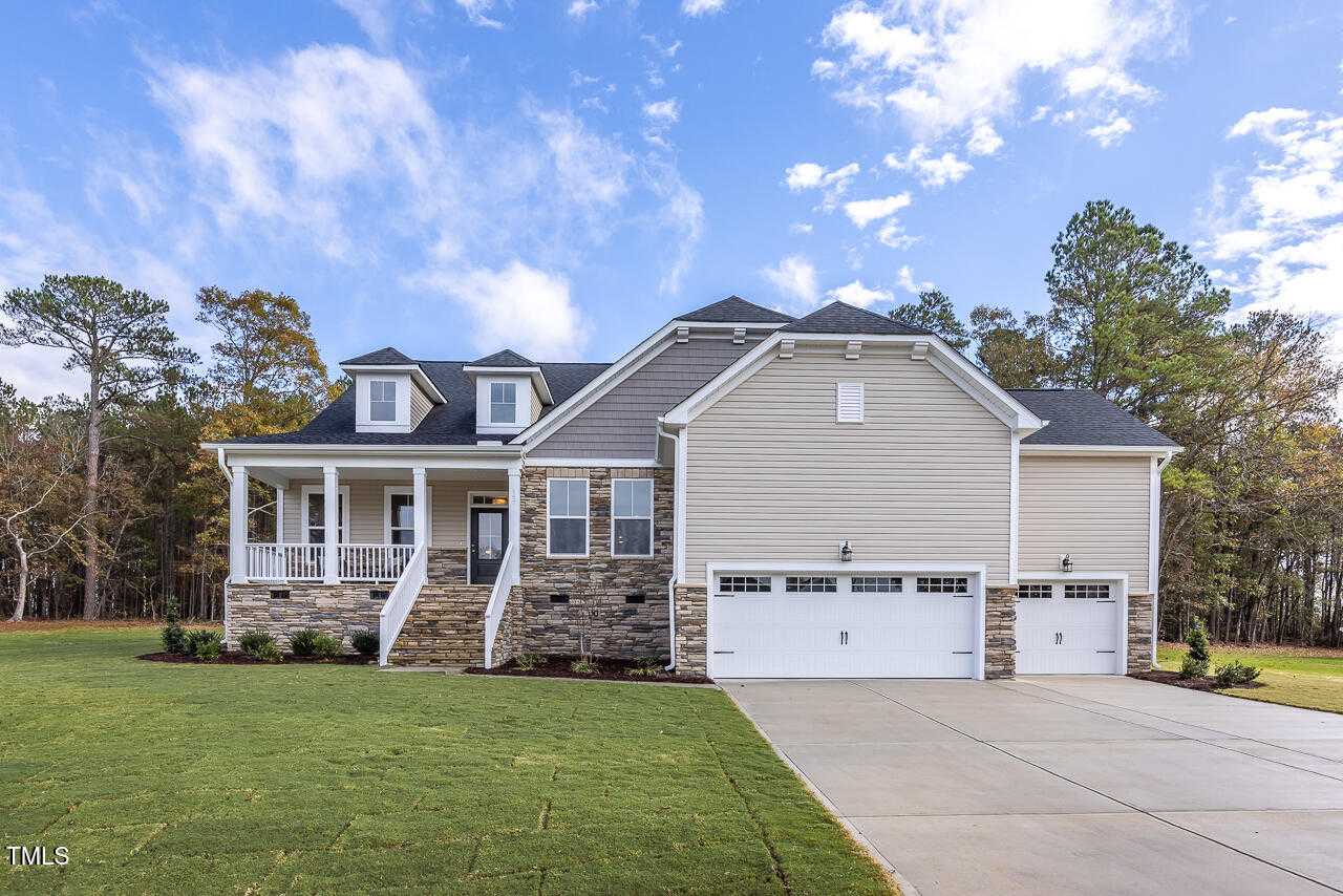 Modern gray Craftsman home with covered front porch, 3-car garage, blue shutters, and lush landscaping in Tobacco Road, Angier, NC