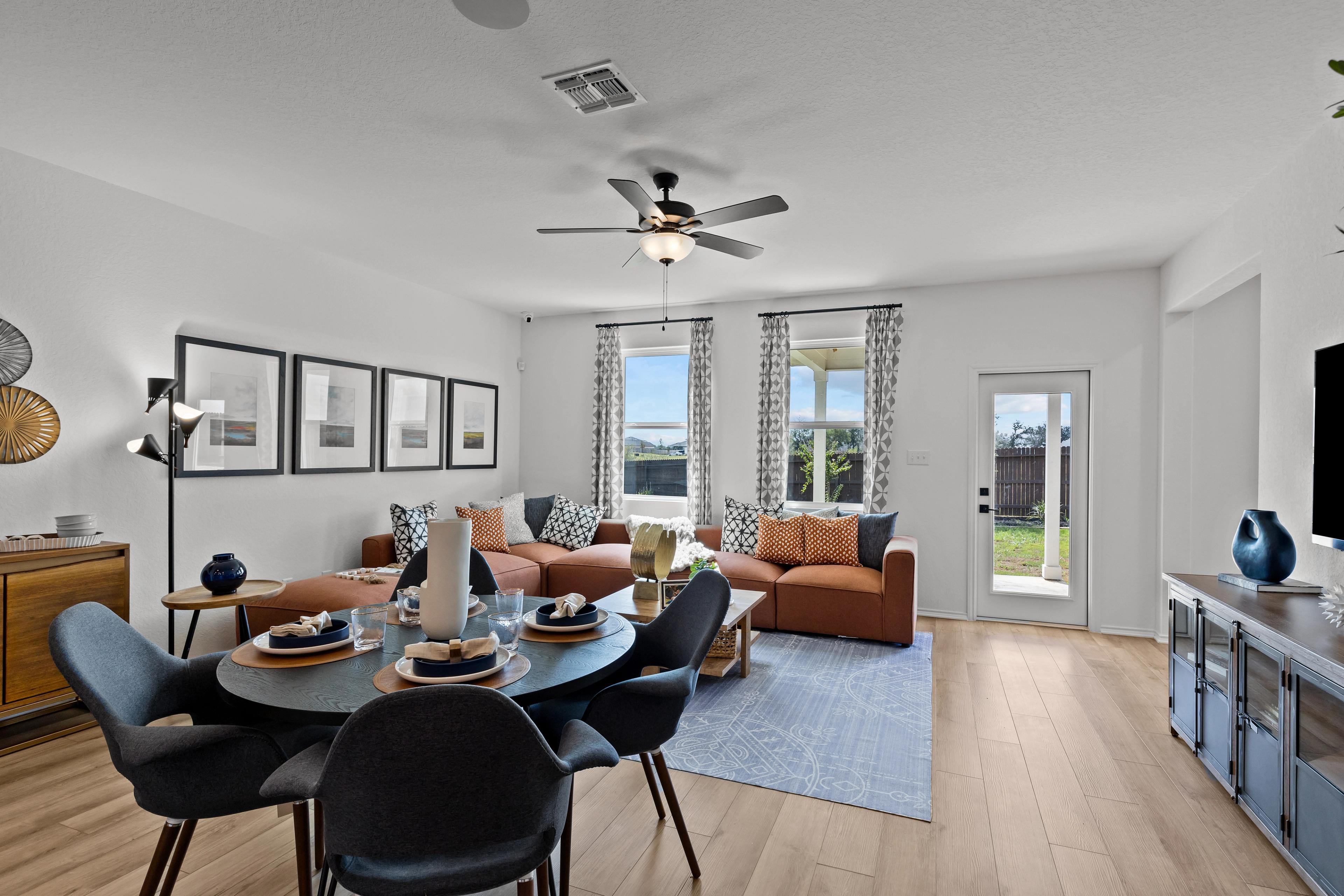 Spacious open-concept living and dining area in The Douglas F with round wooden table, orange sofa, ceiling fan, and large windows to backyard
