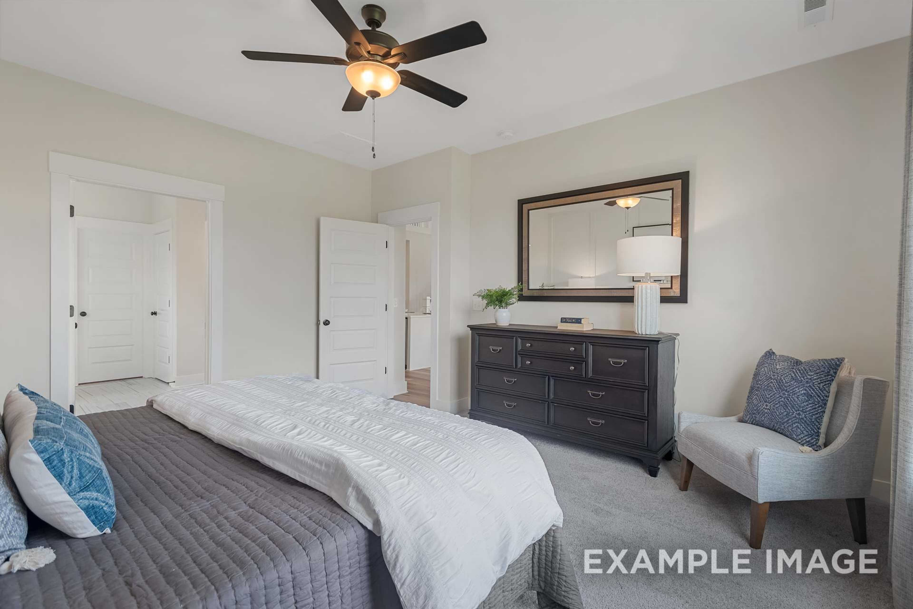Spacious master bedroom in The Ridgeport B with king bed, dark wood dresser, ornate mirror, accent chair, and beige walls