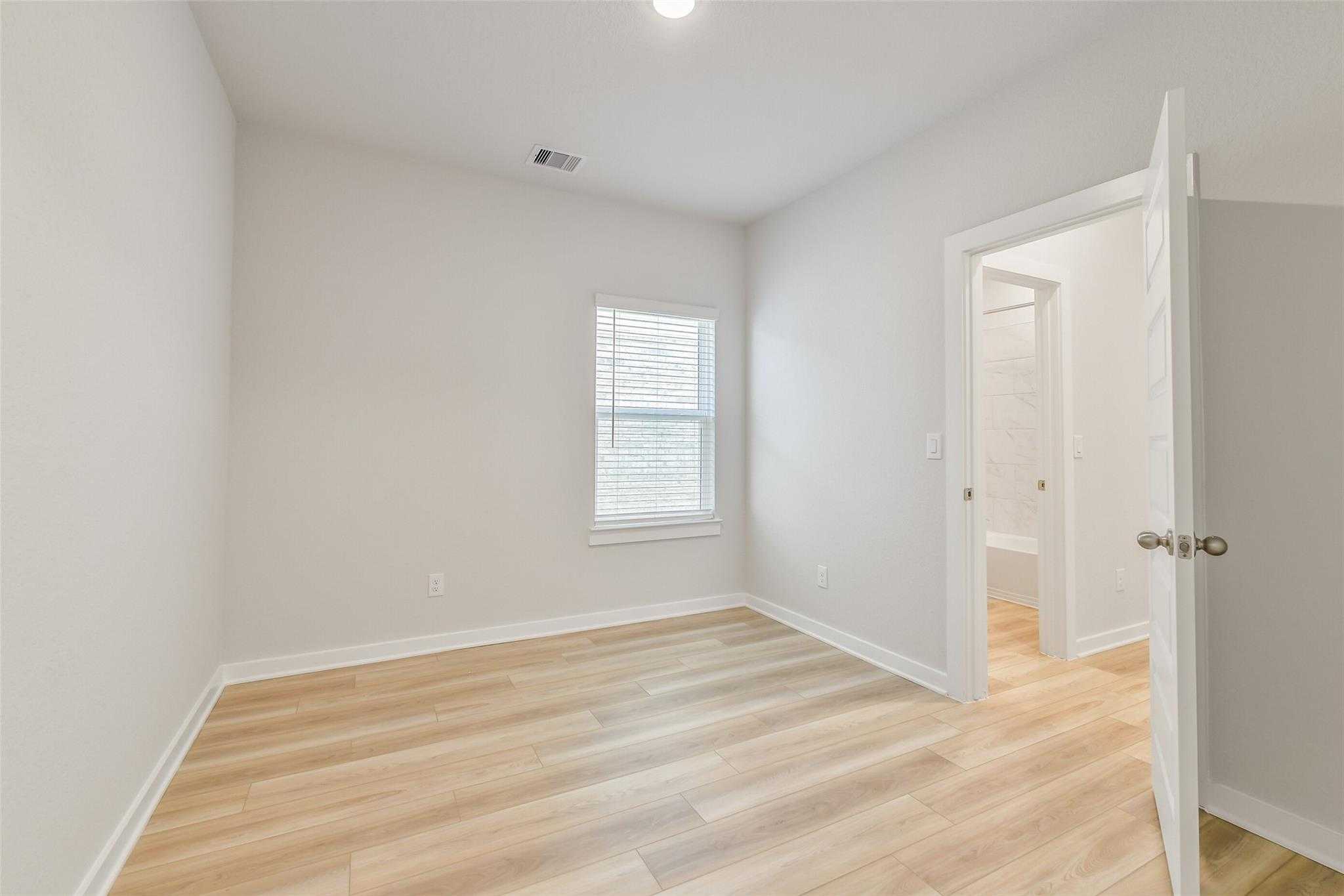 Bright secondary bedroom with light wood floors, white walls, open bathroom door and window blinds in Davidson Homes Rio Grande H, Magnolia Texas