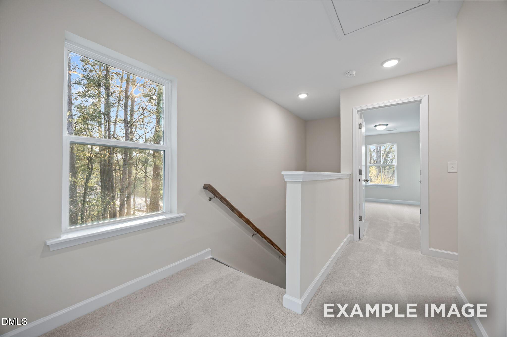 Sunlit upstairs hallway with large windows overlooking trees, wood-railed staircase, and carpeted floors in Davidson Homes The Avery, Knightdale, NC