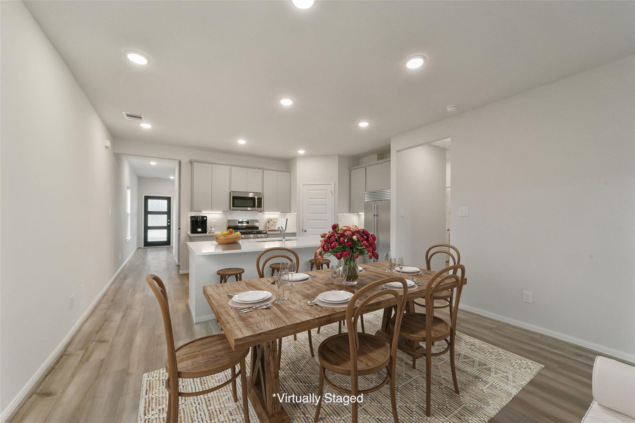 Open-concept dining area with wooden table, chairs, and red roses adjacent to white kitchen in San Marcos E home, Beasley, Texas