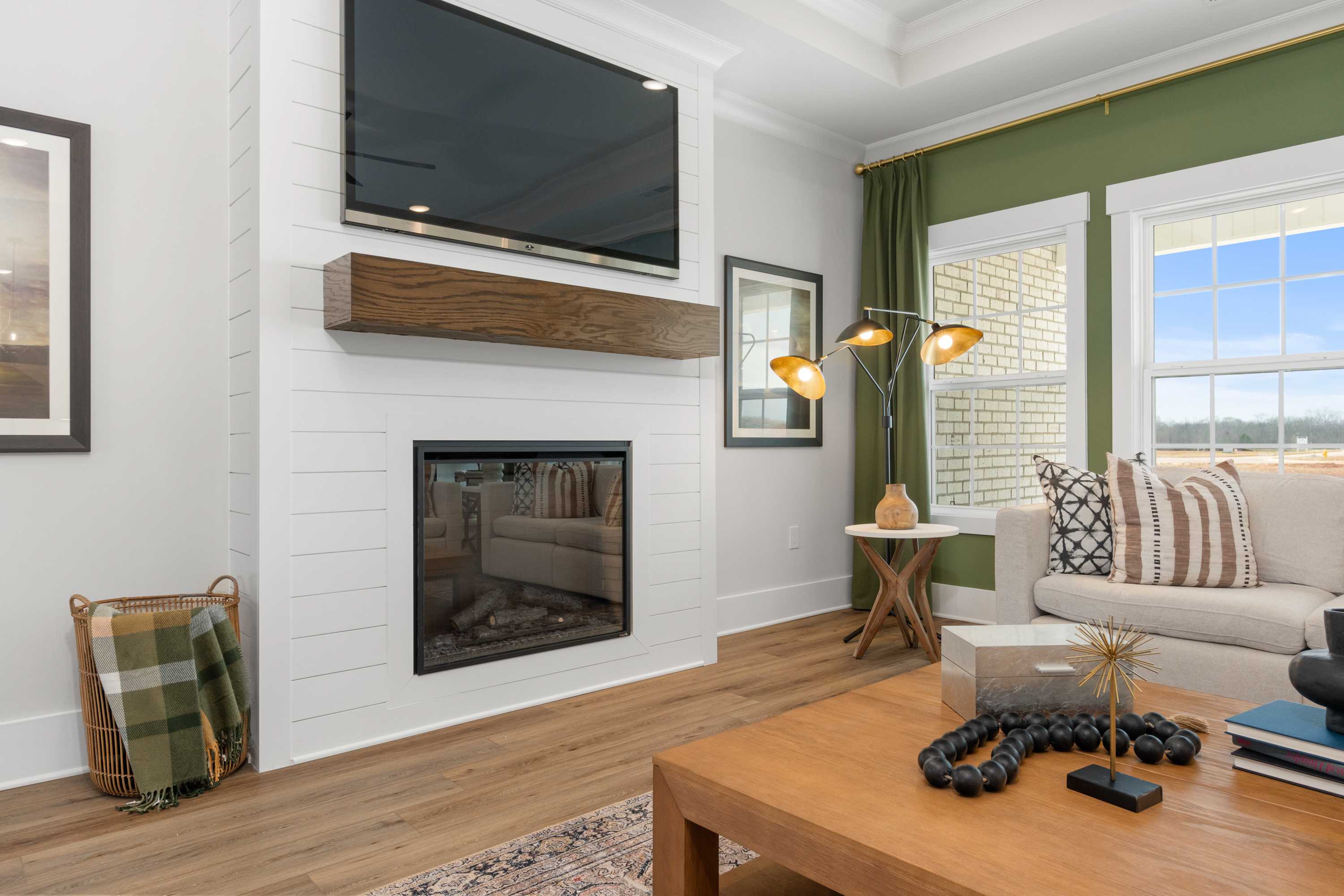 Cozy living room in Briercreek featuring white brick fireplace, mounted TV, beige sofa, hardwood floors, and green-curtained windows in Meridianville, Alabama