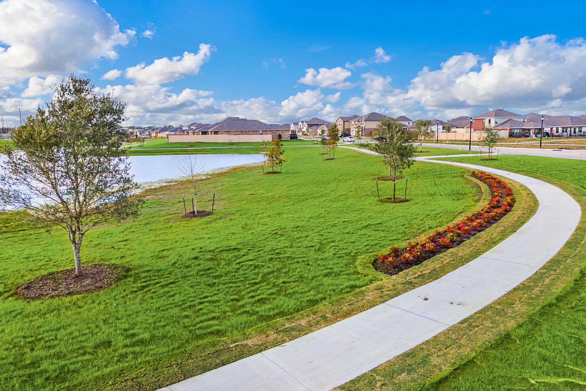 Serene pondside walking path lined with red flowers at Sierra Vista in Rosharon Texas, lush green lawns and modern homes