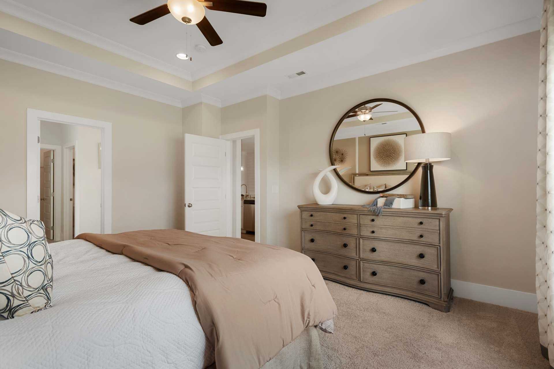 Spacious bedroom at Flint Meadows in New Market Alabama with beige bedding, wooden dresser, round mirror, and ceiling fan