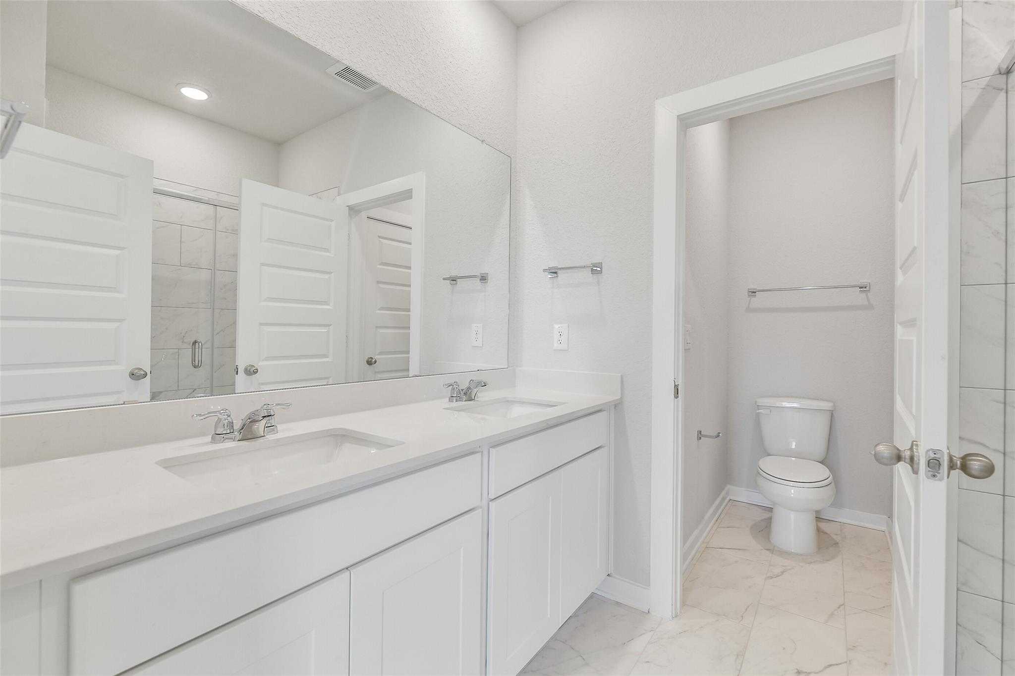 Elegant master bathroom with double white vanity, quartz counters, large mirror, and separate toilet in Davidson Homes The Colorado G, Magnolia, Texas