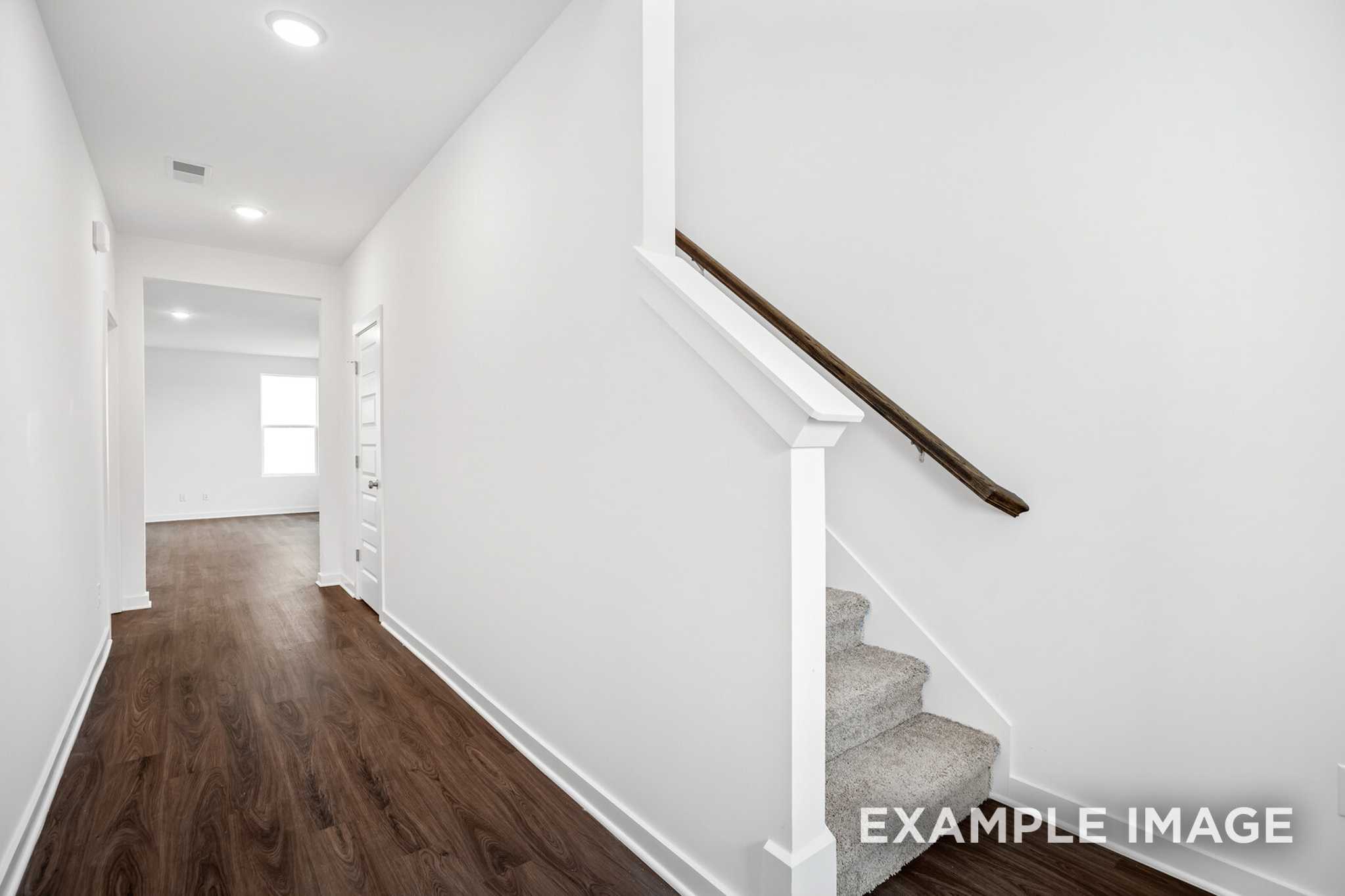 Bright hallway in The Grayson C home with dark hardwood floors, white walls, and carpeted staircase to upper master suite