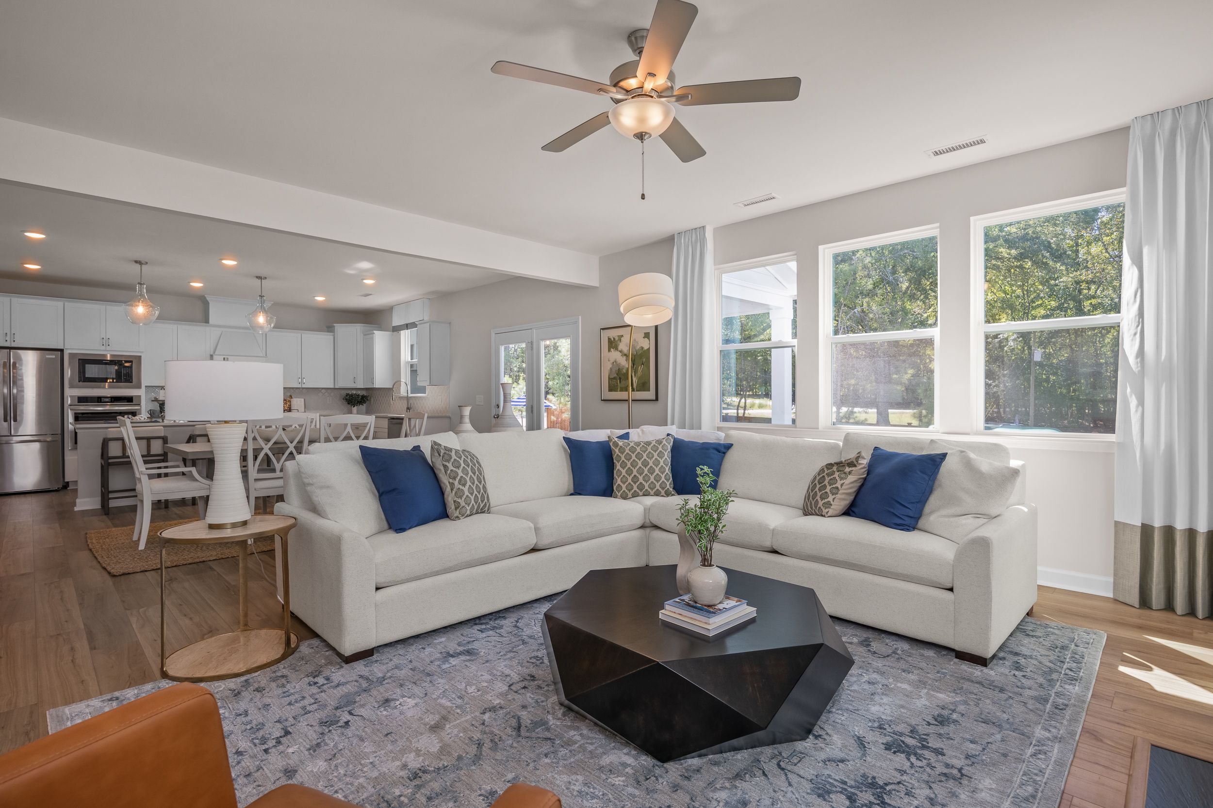 Open-concept living room at Enclave at Belmont in Belmont NC with L-shaped sofa, hardwood floors, blue rug, and adjacent white kitchen