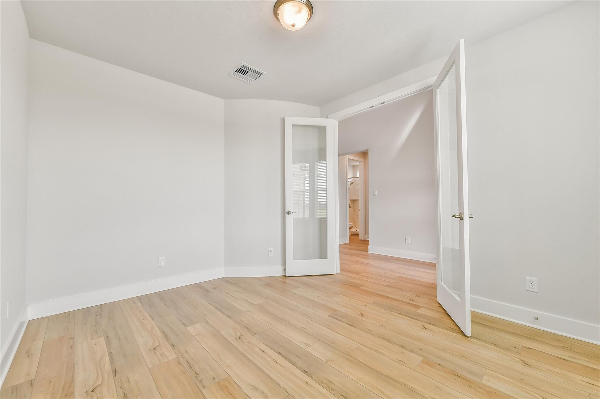 Bright bedroom with light oak hardwood floors, white walls, open glass French doors to en-suite bathroom in Davidson Homes The Victoria A, Lago Mar, Texas City