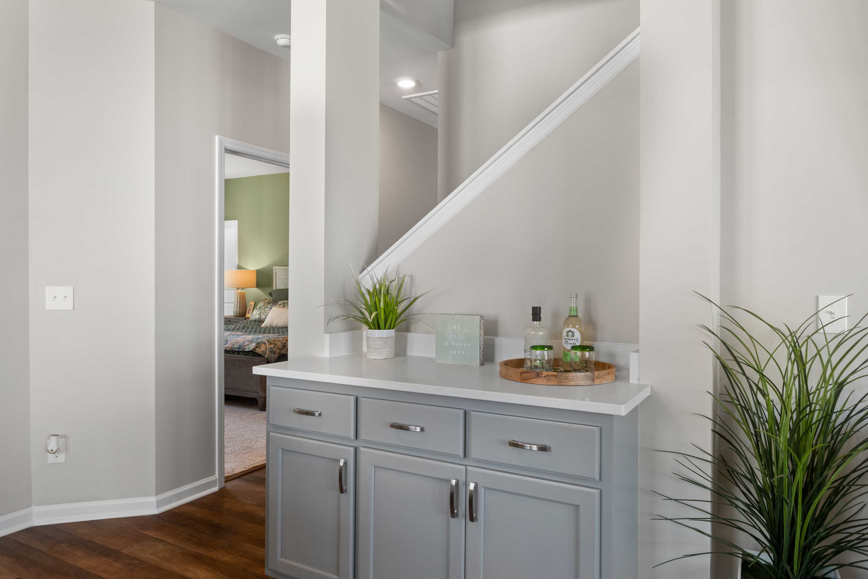 Elegant gray wet bar cabinet in Evergreen Mill home hallway, Madison Alabama, with plants, bottles, and open stairs