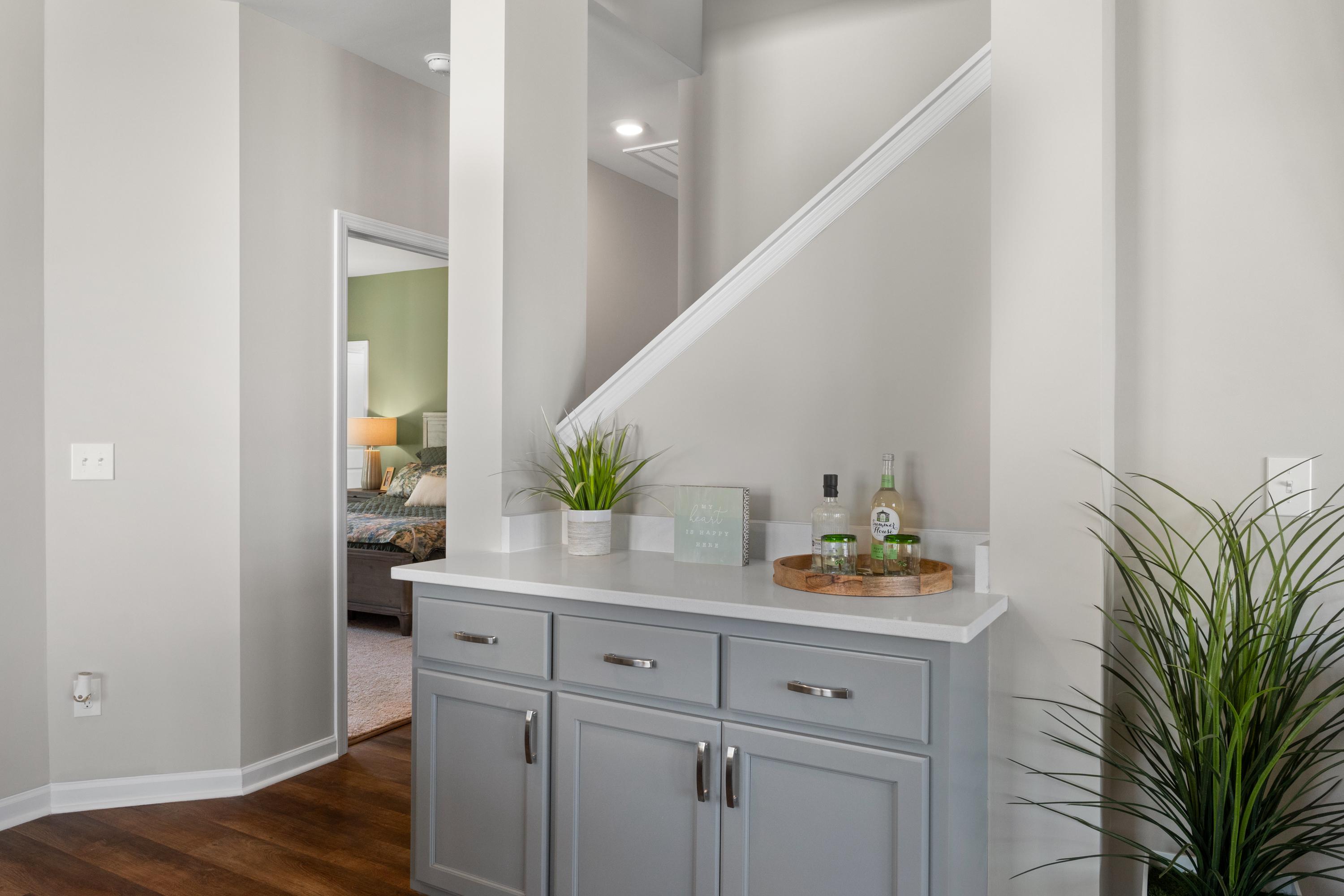 Elegant gray wet bar cabinet in Evergreen Mill home hallway, Madison Alabama, with plants, bottles, and open stairs