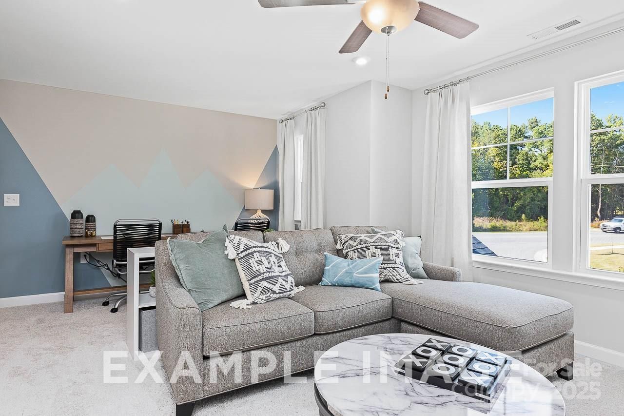 Cozy living room featuring gray L-shaped sectional, geometric blue-gray accent wall, ceiling fan, and wooded views in Davidson Homes The Hemlock A, Belmont, NC