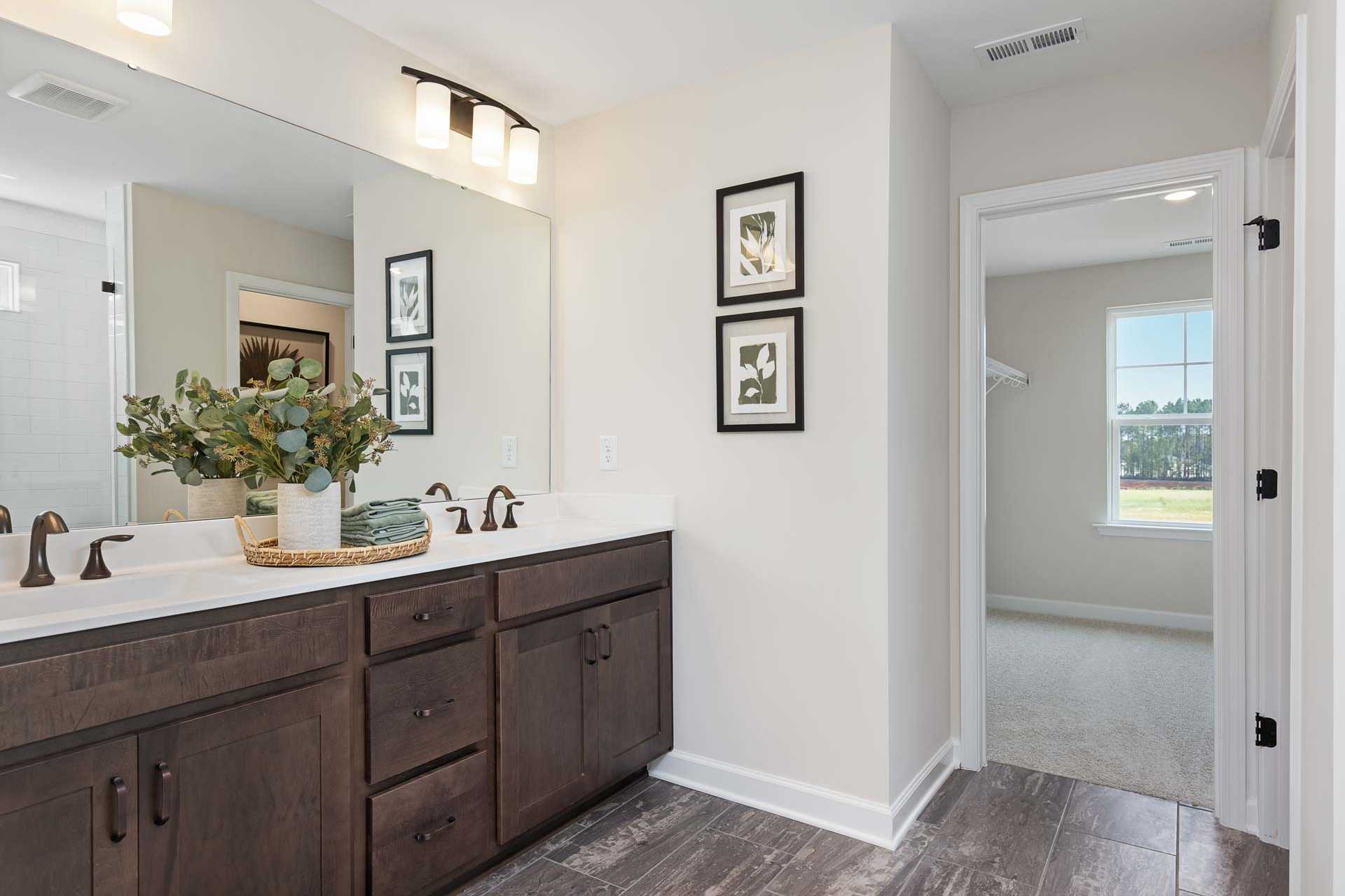 Spacious primary bathroom at Sierra Heights in Clayton NC with double vanity, eucalyptus vase, framed art, and adjacent bedroom view