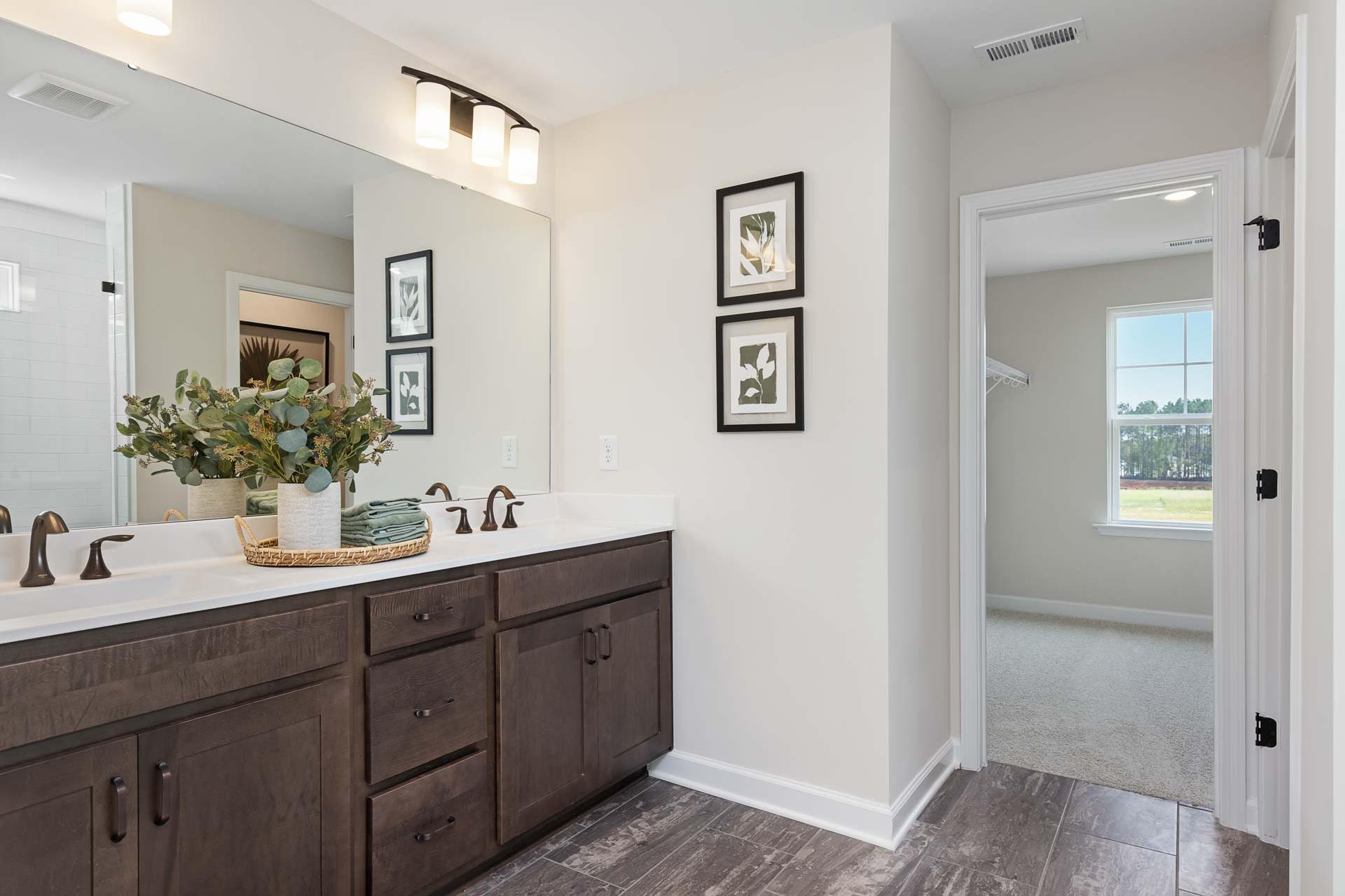 Spacious primary bathroom at Sierra Heights in Clayton NC with double vanity, eucalyptus vase, framed art, and adjacent bedroom view