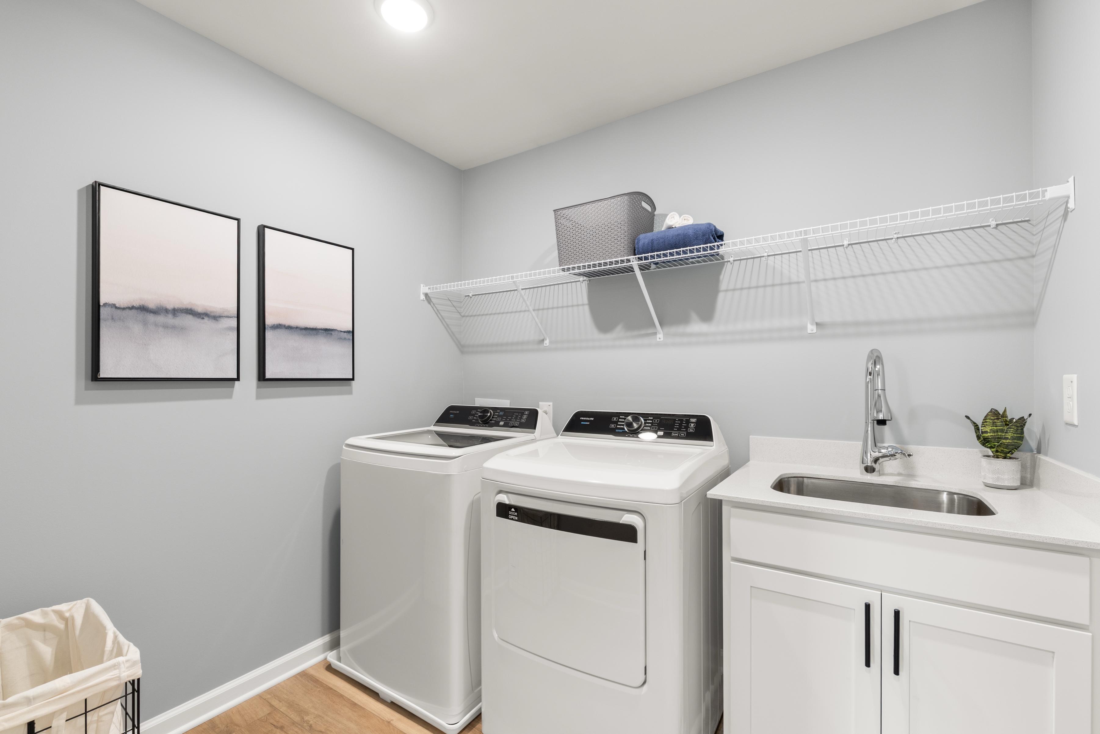 Spacious laundry room at The Bluffs in Canton, Georgia by Davidson Homes with white washers, dryers, utility sink, wire shelving, and abstract wall art