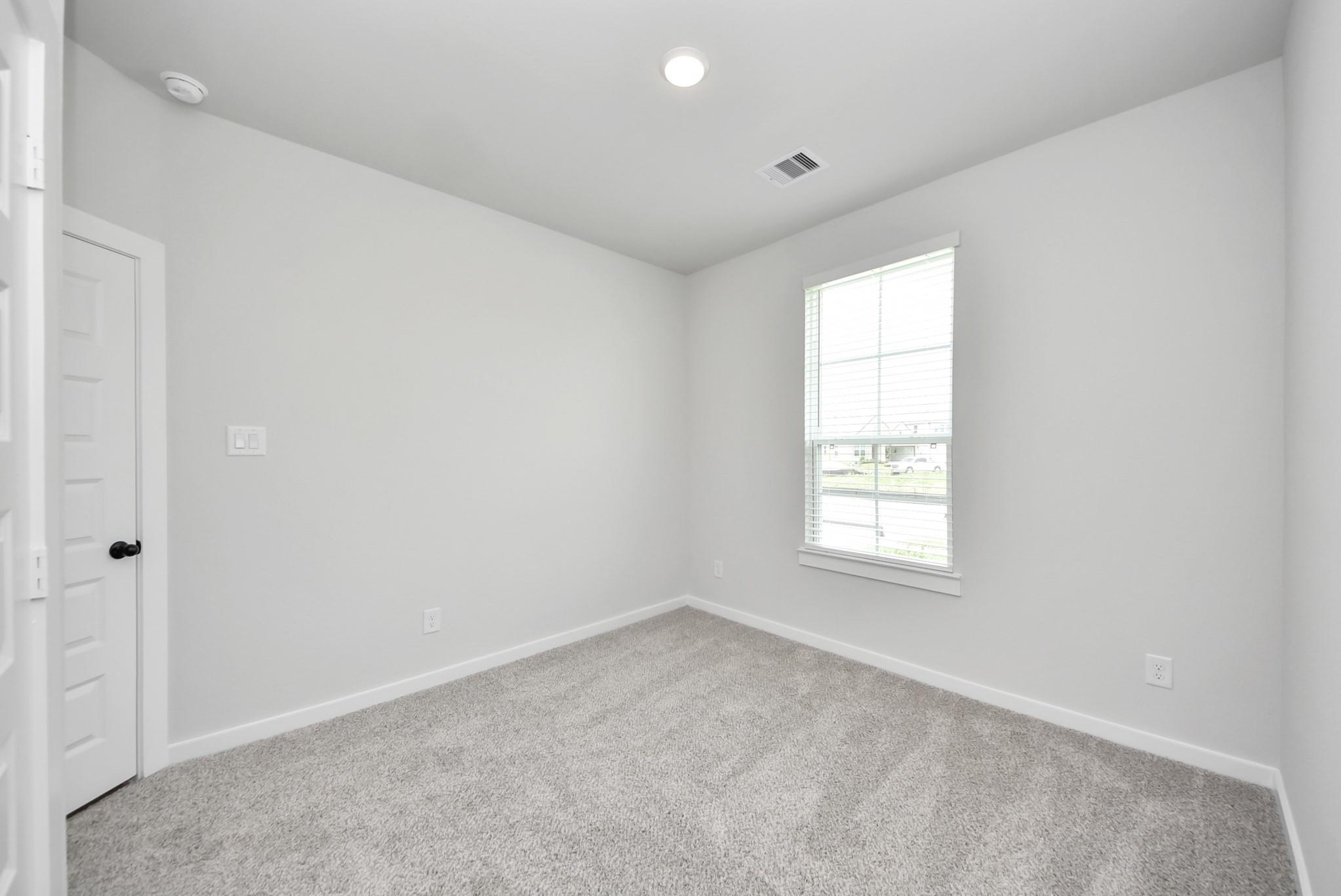 Bright empty bedroom with gray carpet, white walls, and window with blinds in Davidson Homes The Brookside F plan, Crosby, Texas