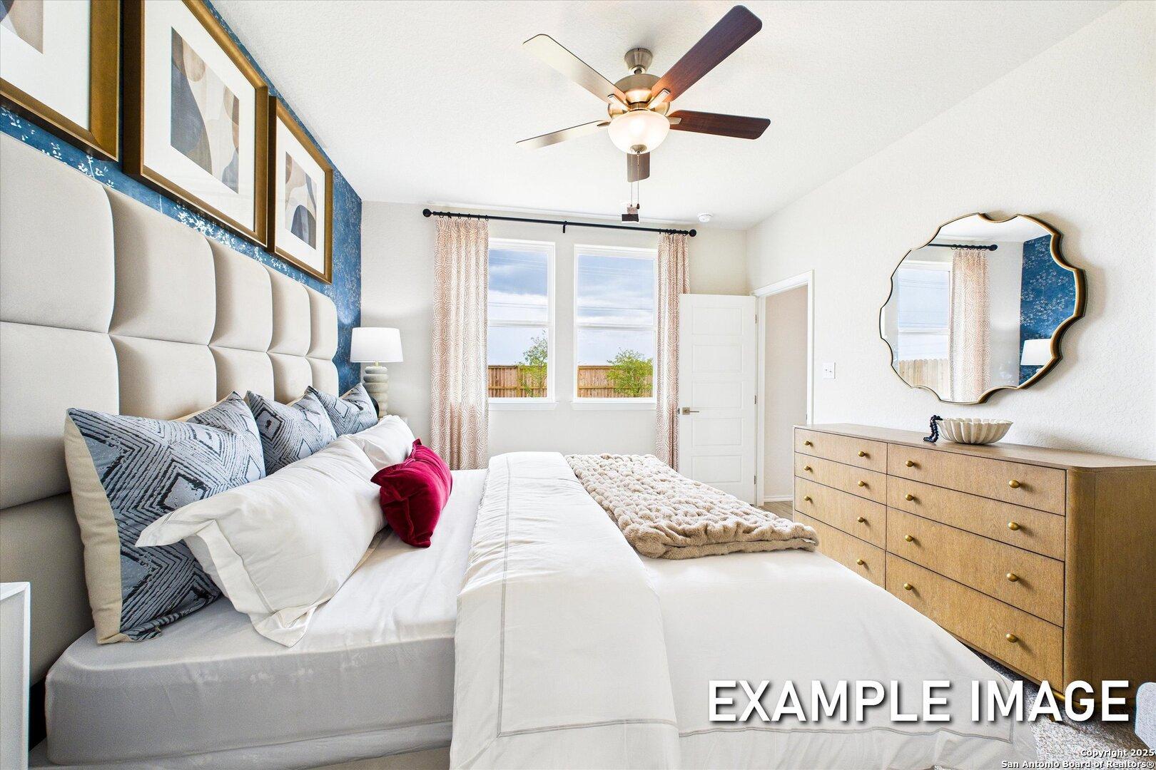 Elegant master bedroom with tufted beige headboard, white bedding, blue pillows, wooden dresser, ceiling fan in Davidson Homes The Brazos C, San Antonio