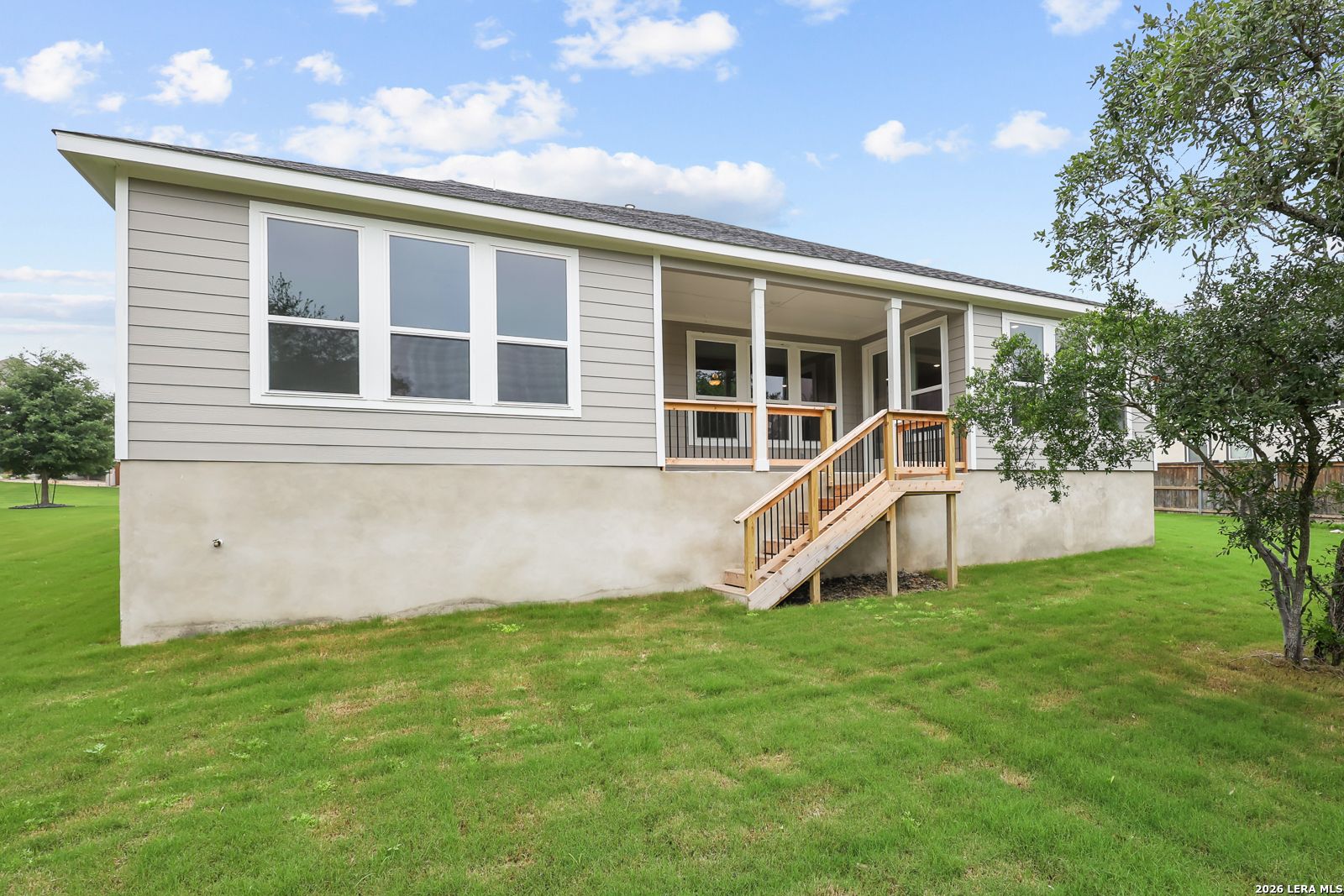 Two-story gray-sided home with covered porch, large windows, wooden deck stairs, and lush green yard in Potranco Oaks, Castroville, Texas