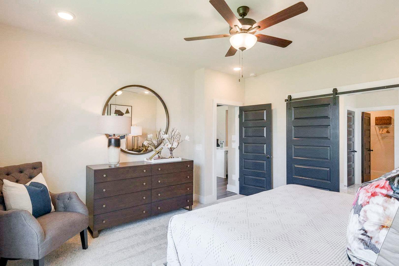 Spacious master bedroom at The Meadows in Smyrna TN featuring barn doors, ceiling fan, and tufted armchair
