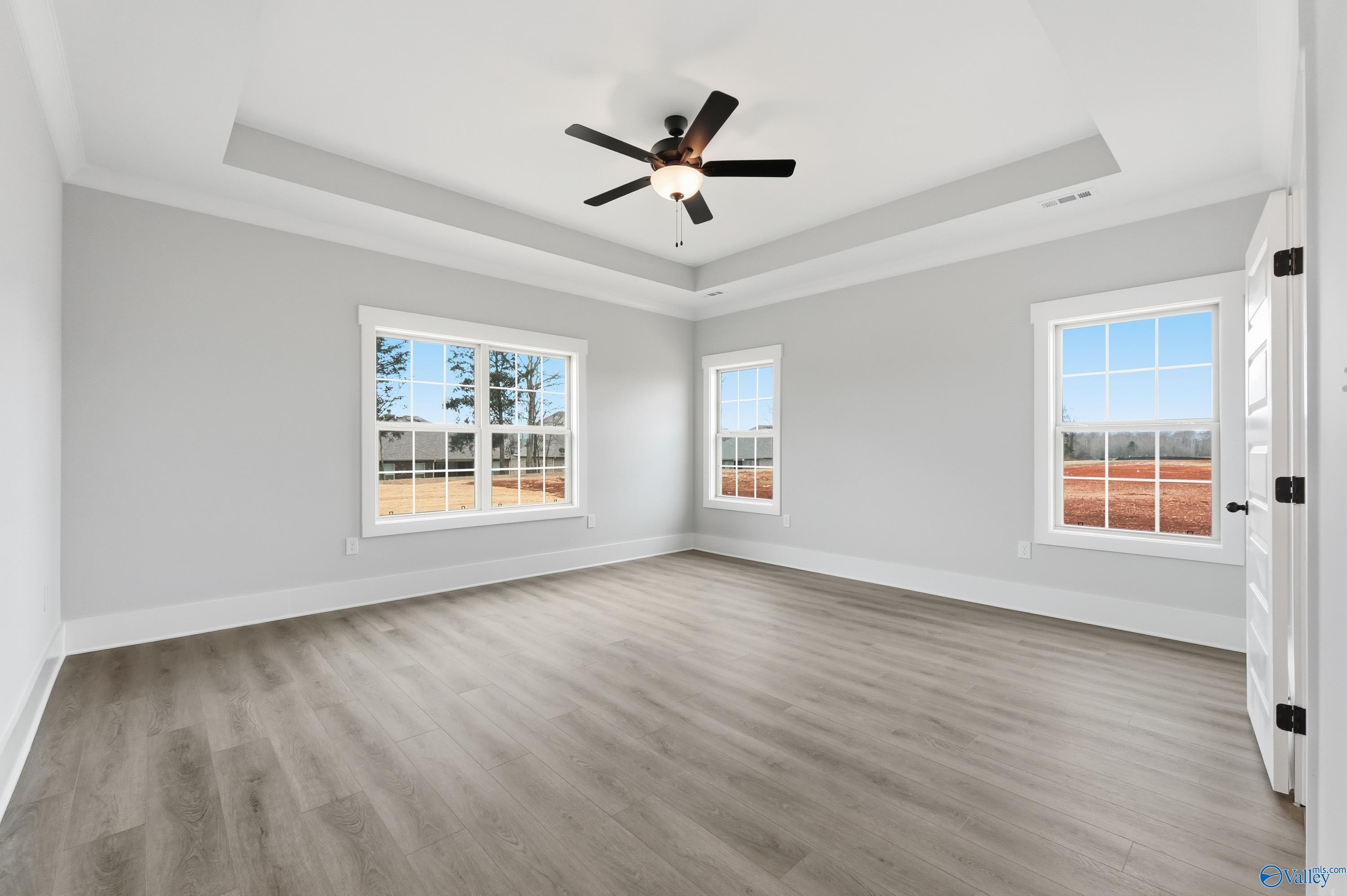 Bright master bedroom with tray ceiling, ceiling fan, large windows overlooking trees, and hardwood floors in Davidson Homes The Finleigh, Meridianville, AL