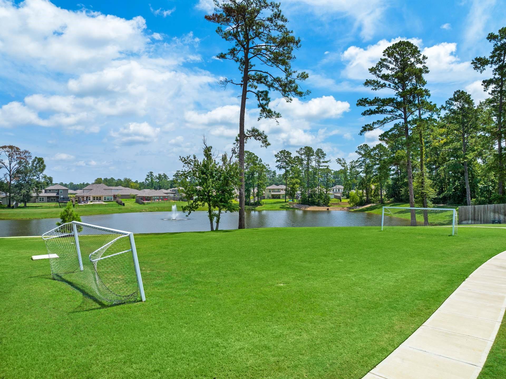 Scenic pondside soccer field with white goals on lush green grass, pine trees, and homes in Lakes at Black Oak, Magnolia, Texas