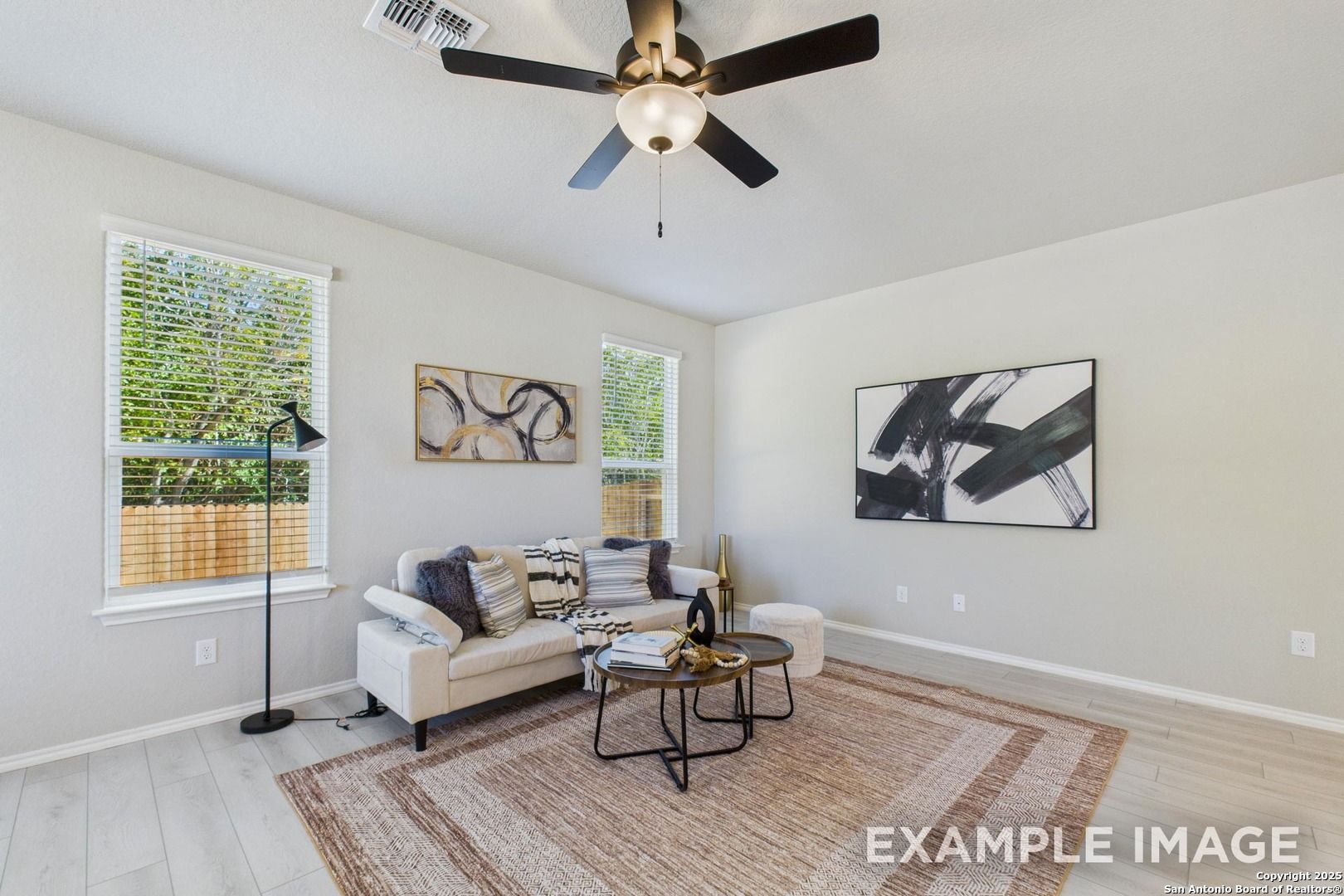 Cozy living room with beige sofa, ceiling fan, abstract art, and tree-view windows in Davidson Homes The Charlotte A, San Antonio
