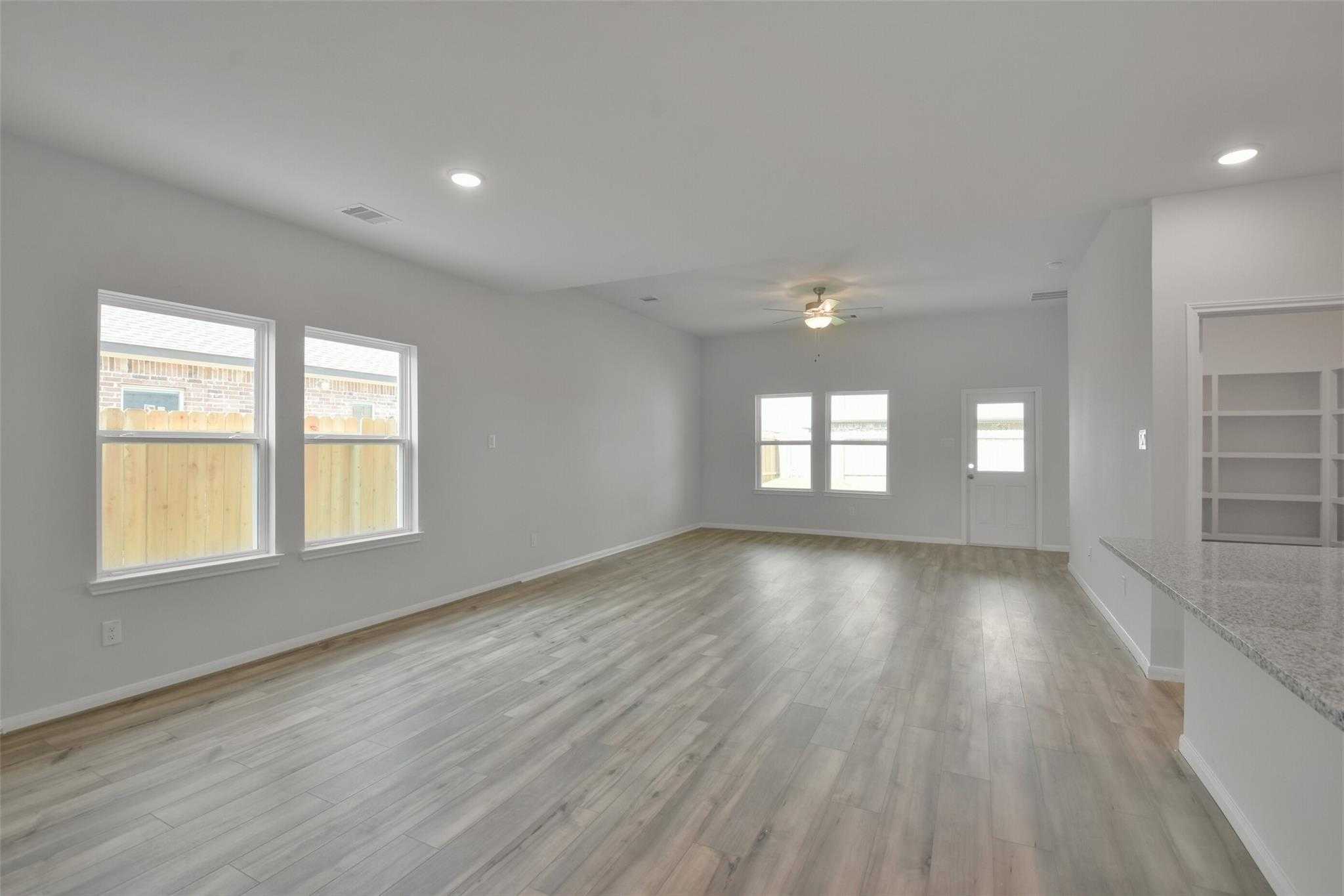Spacious open living room with hardwood floors, large windows, ceiling fan, and adjacent granite kitchen in Davidson Homes The Tierra A, Dayton, Texas