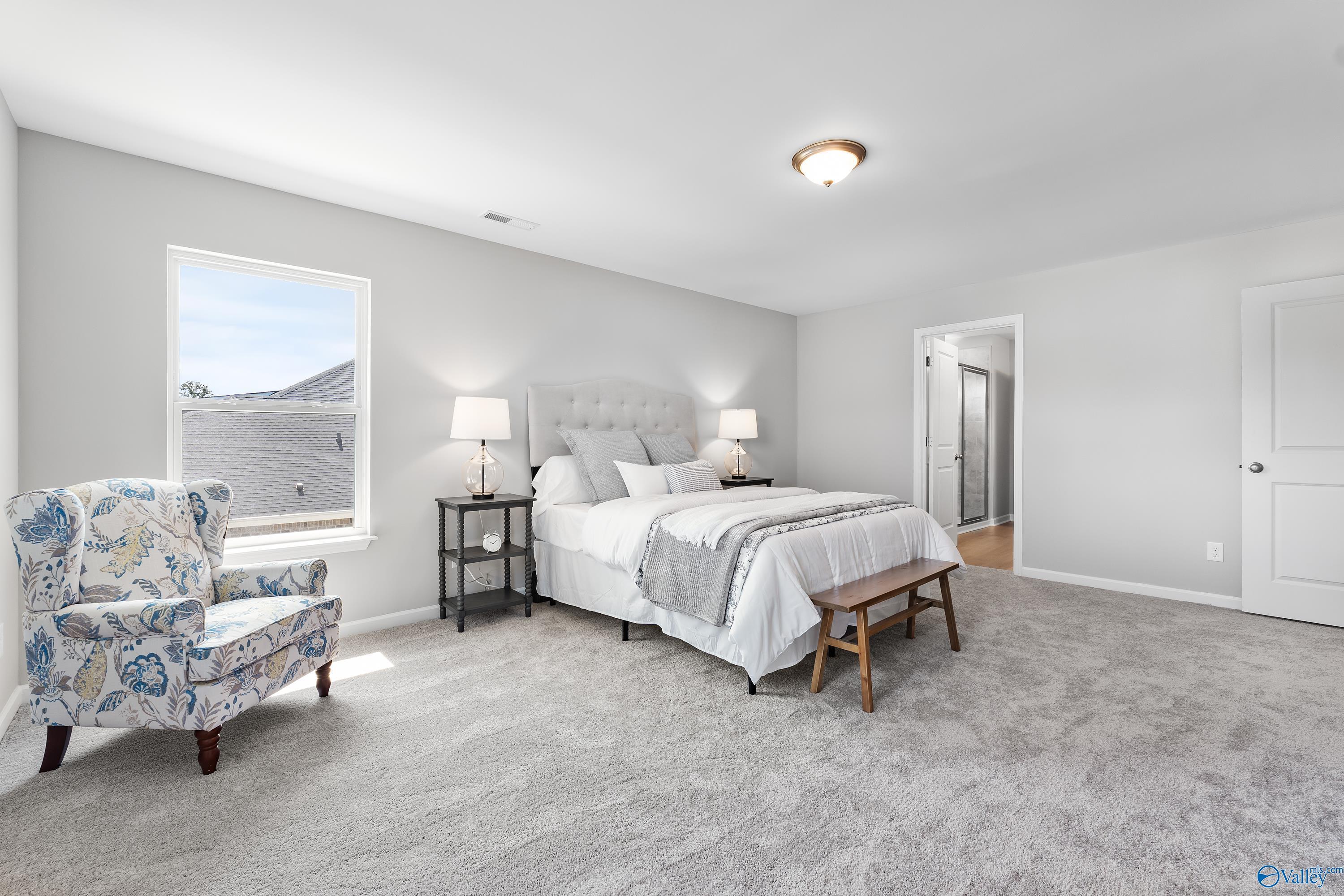 Serene master bedroom with king bed, tufted gray headboard, floral armchair, and natural light in Davidson Homes The Charm, Huntsville AL