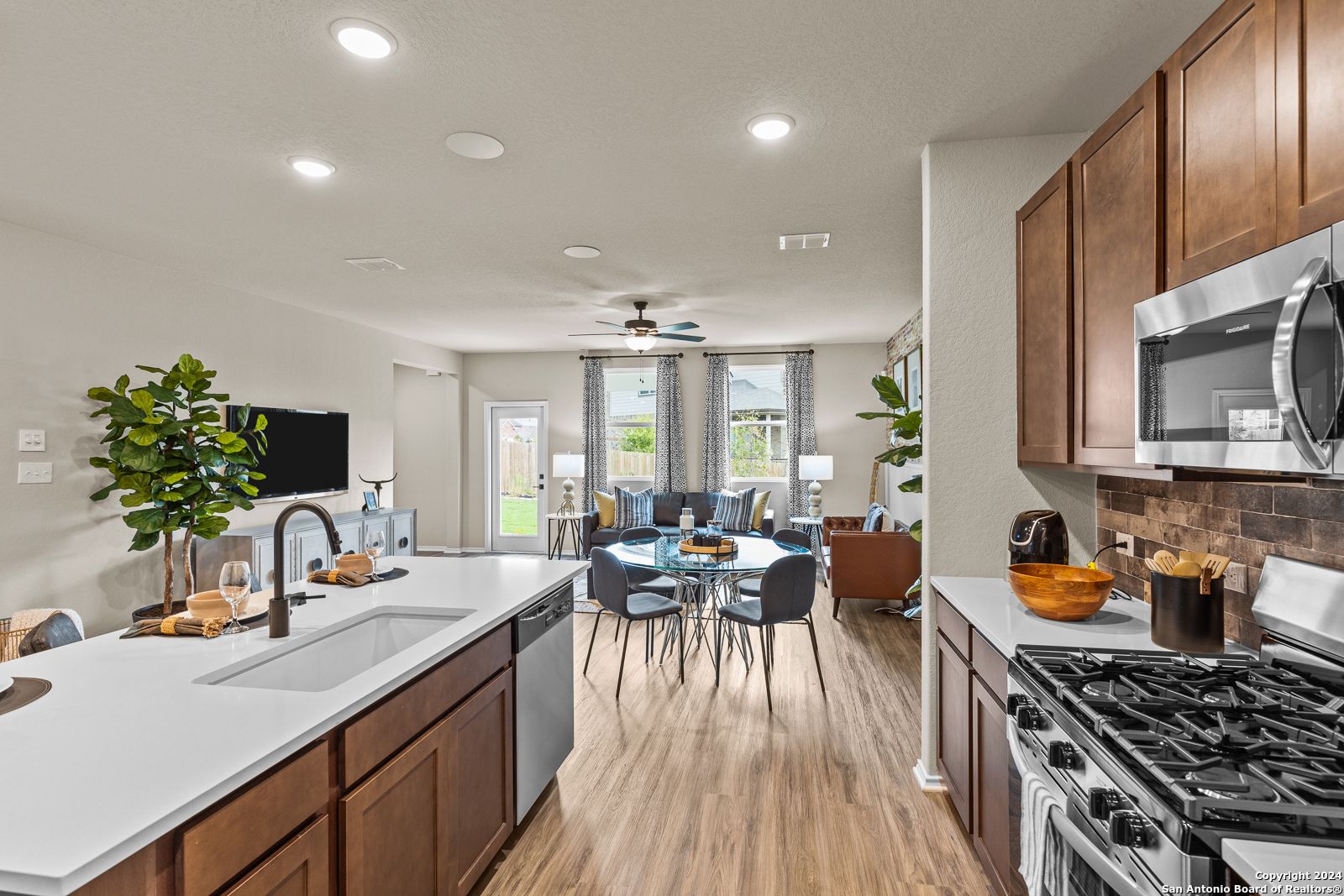 Open-concept kitchen with wood cabinets, stainless appliances, island sink, and adjacent living-dining area in The Douglas F, Bricewood, San Antonio