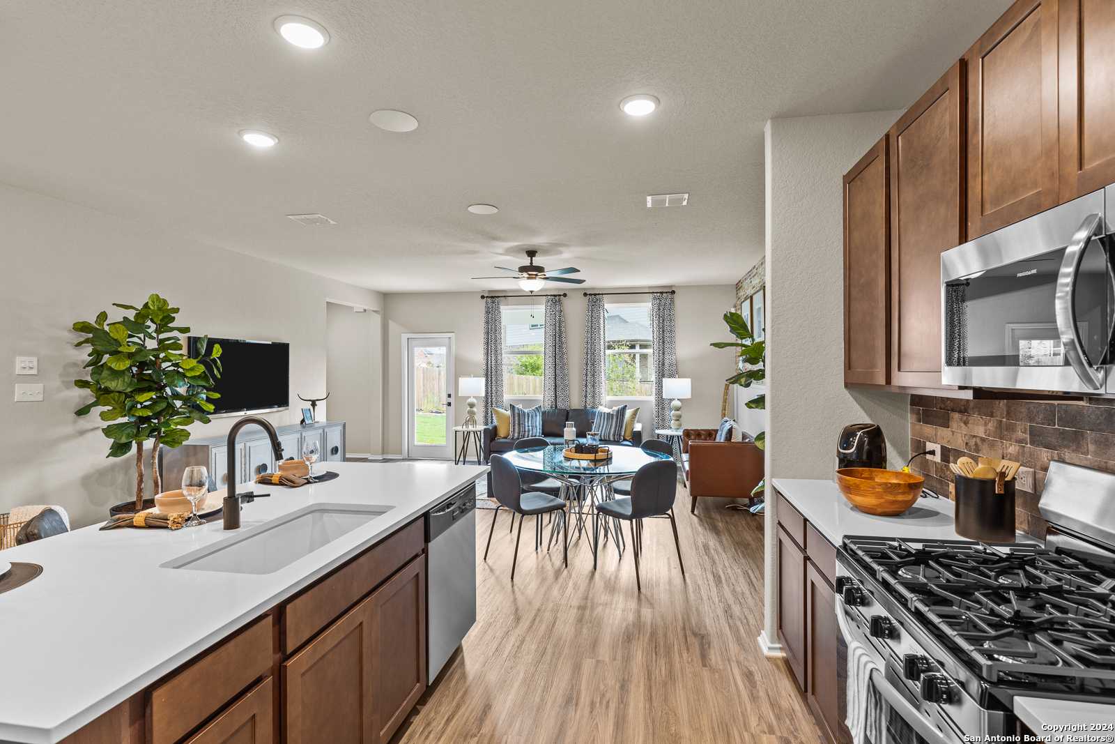 Open-concept kitchen with wood cabinets, stainless appliances, island sink, and adjacent living-dining area in The Douglas F, Bricewood, San Antonio