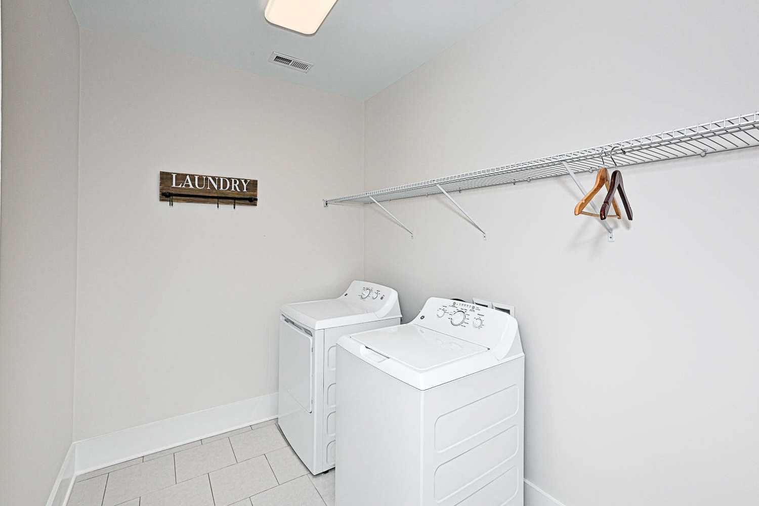 Spacious laundry room in The Finleigh featuring white washer-dryer set, wire shelving, and wooden sign