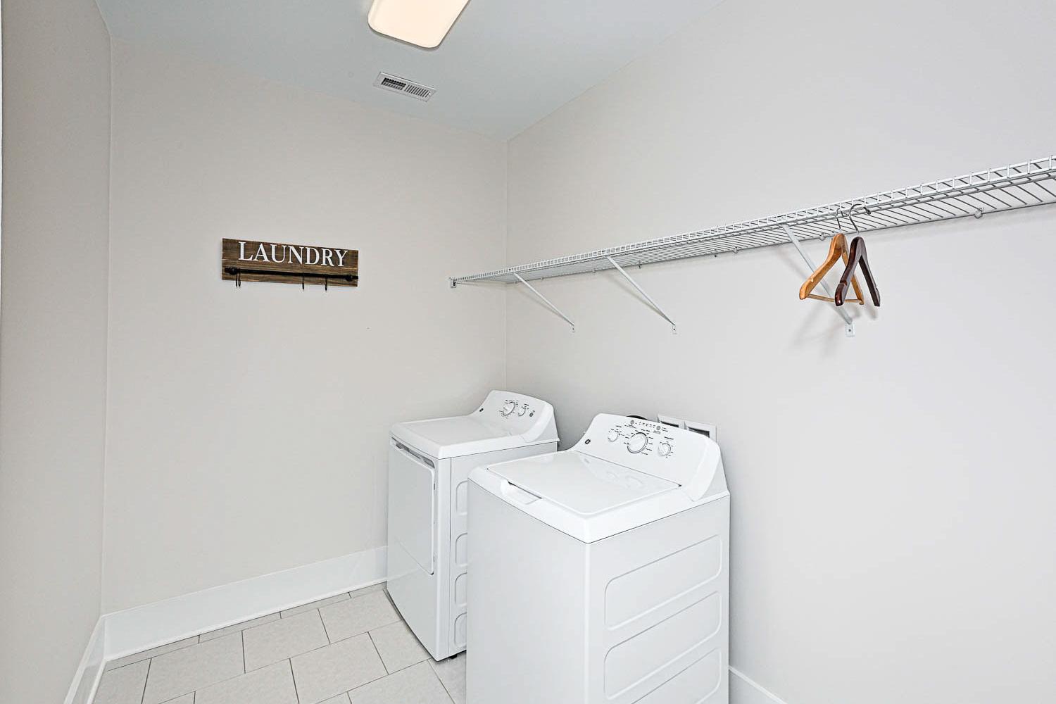 Spacious laundry room in The Finleigh featuring white washer-dryer set, wire shelving, and wooden sign