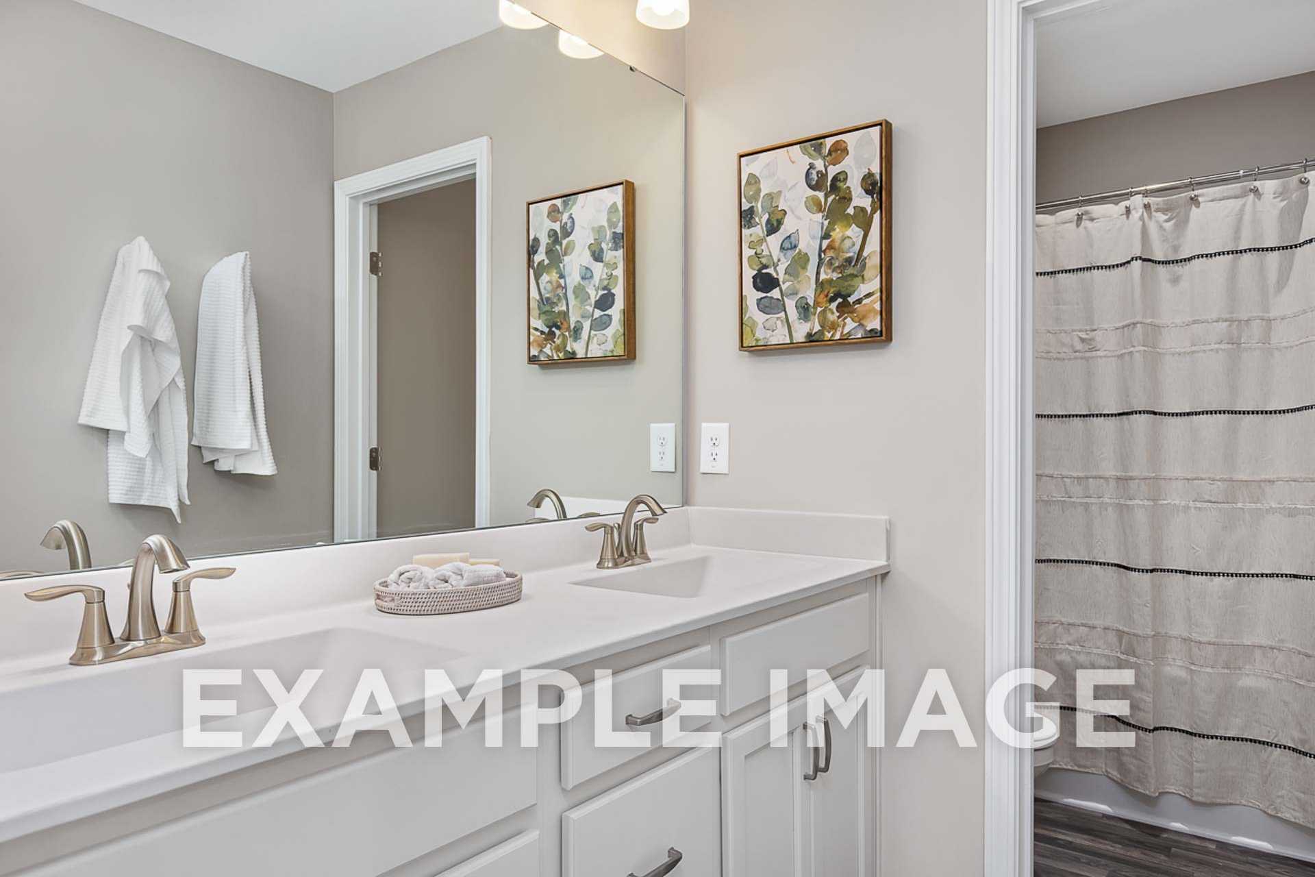 Spacious master bathroom in The Ash E with double vanity, gold faucets, gray shower curtain, and leaf artwork