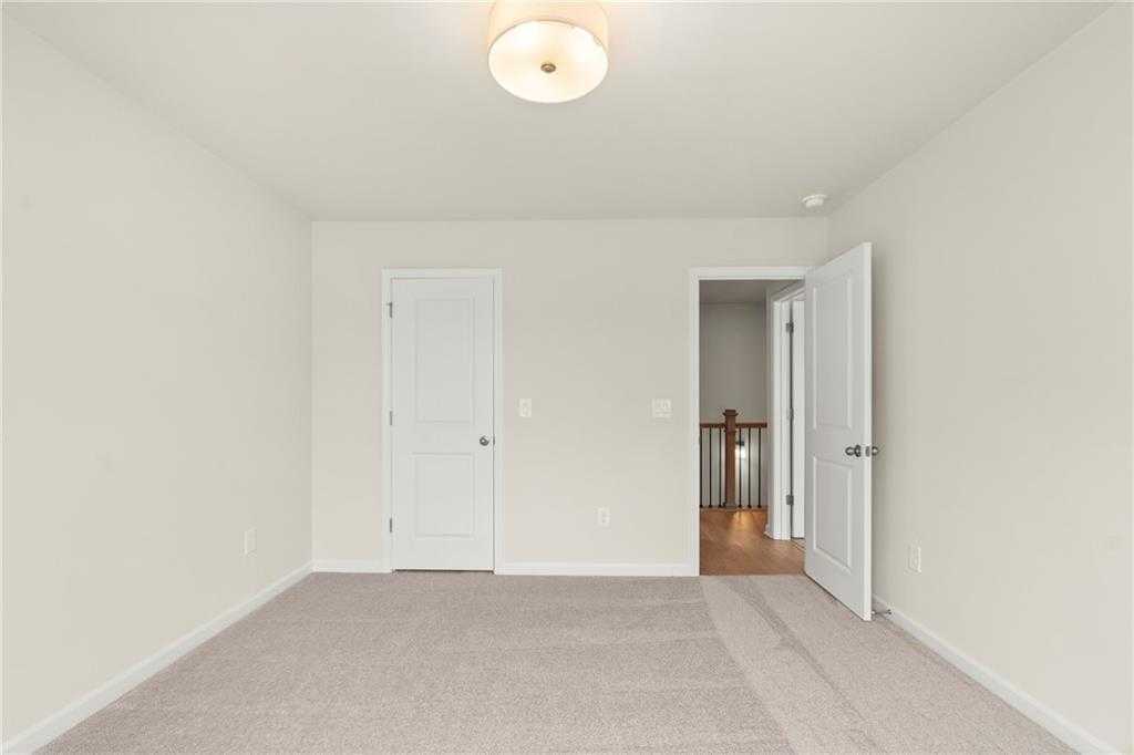 Spacious empty bedroom with beige walls, carpeted floor, and open door to hallway stairs in Davidson Homes The Cary A, Kennesaw, Georgia
