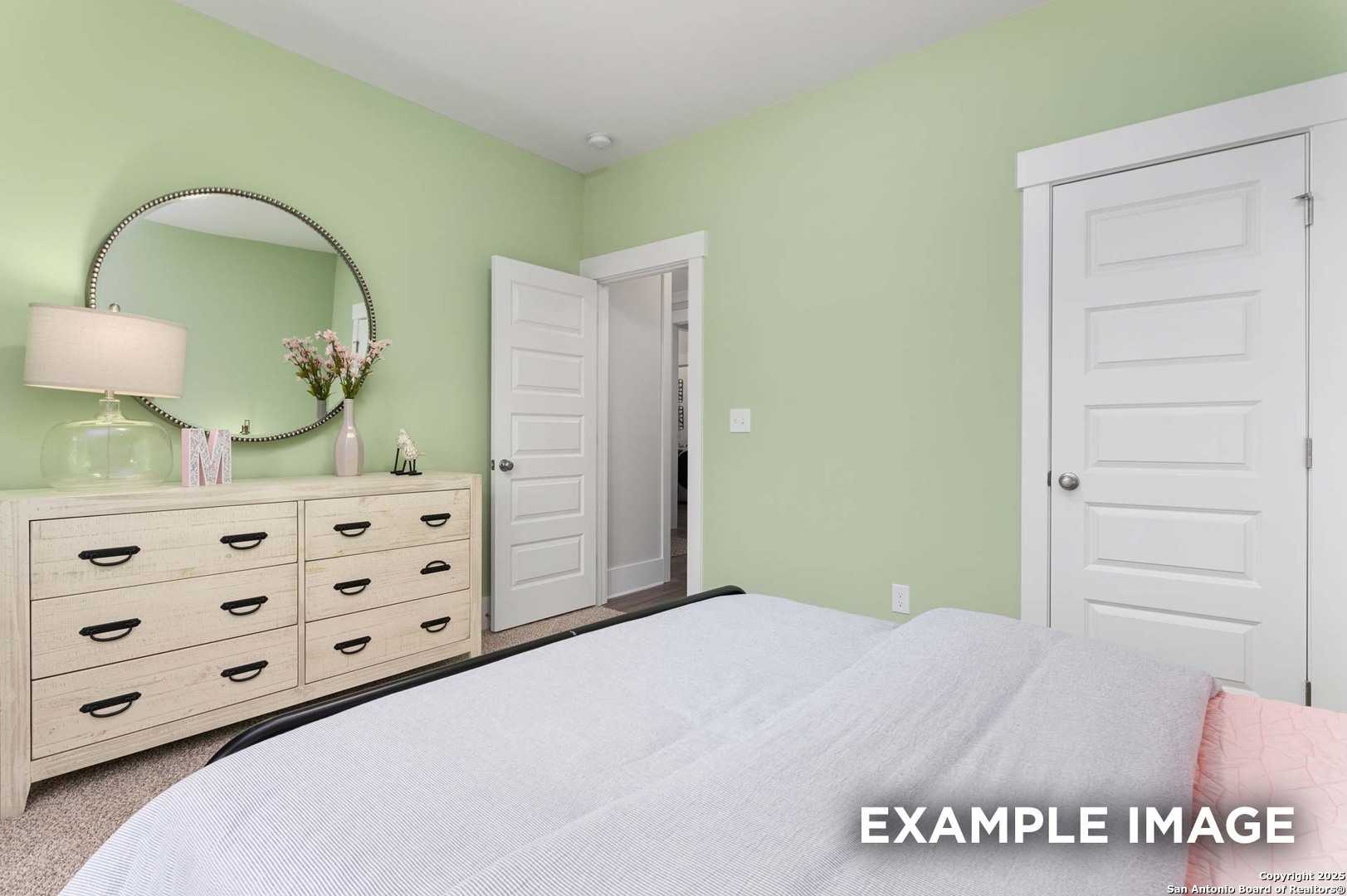 Cozy sage green bedroom with white bed, wooden dresser, round mirror, and floral vase in Davidson Homes The Daphne H, Seguin Texas