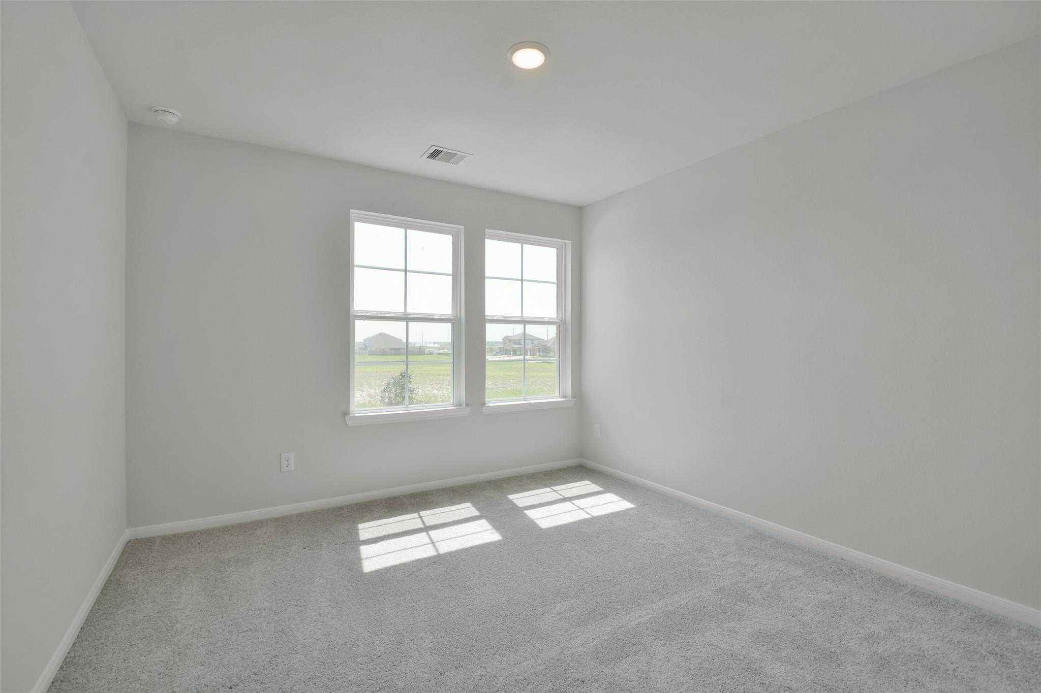 Sunlit empty bedroom with light gray walls, plush carpet, and double windows overlooking fields in Davidson Homes The Tierra A, Dayton, Texas