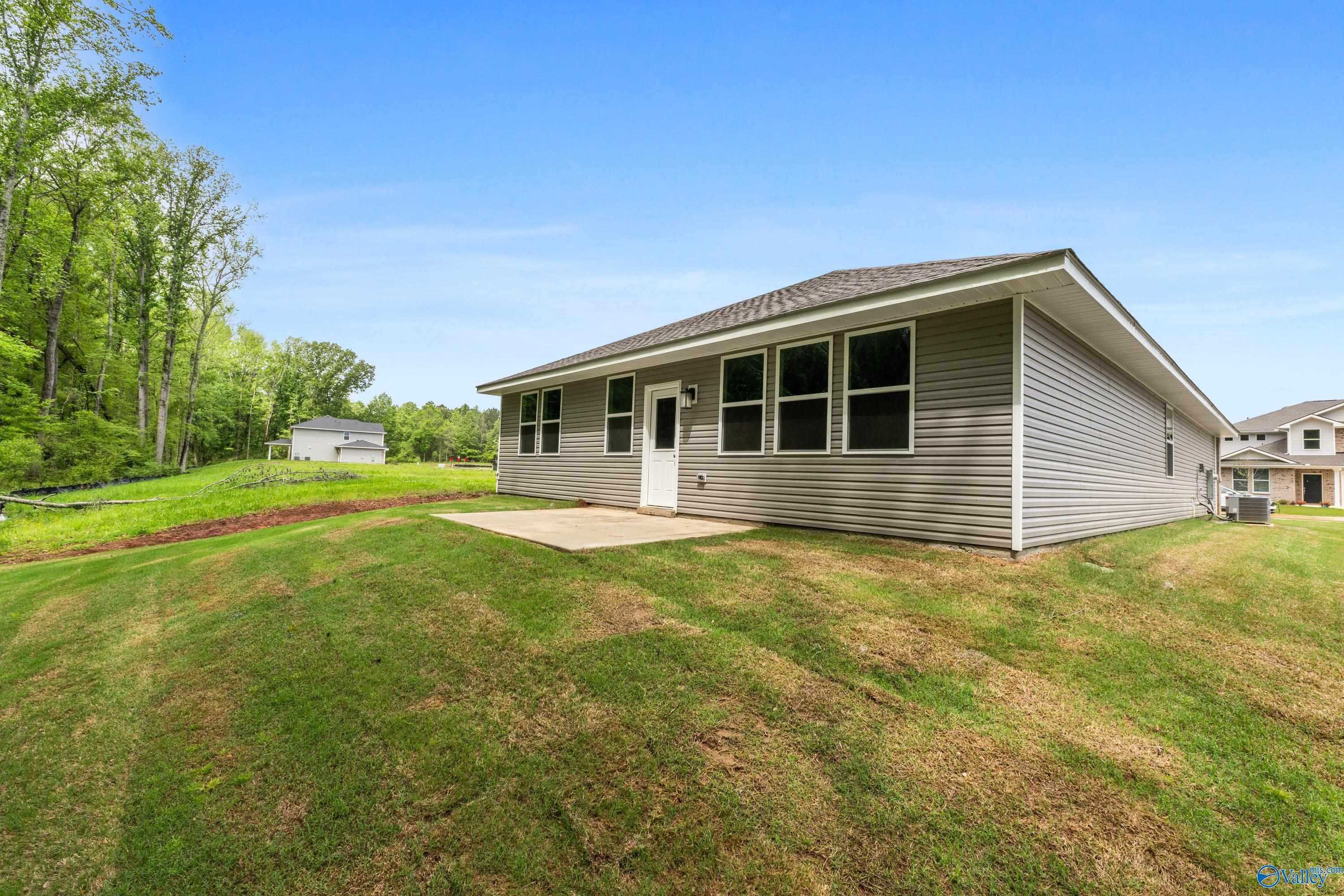 Single-story 4-bedroom home with gray siding, large windows, concrete patio, and lush green yard in Forest Glen, Hazel Green, Alabama - Davidson Homes The Luna