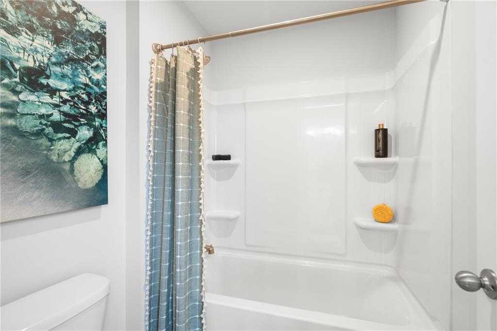 Bright bathroom with white tub, ombre blue-gray shower curtain, bronze rod, shelves holding soap dispenser and loofah, abstract wall art in The Wilmington B, Emerson, Georgia