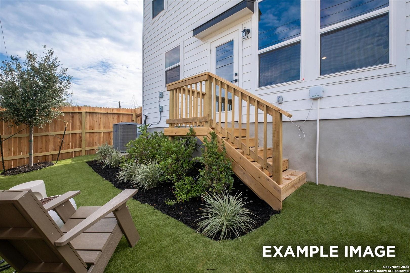 Landscaped backyard with wooden deck stairs to two-story white home, synthetic turf, Adirondack chairs, and privacy fence in Meadows at Oak Creek, San Antonio