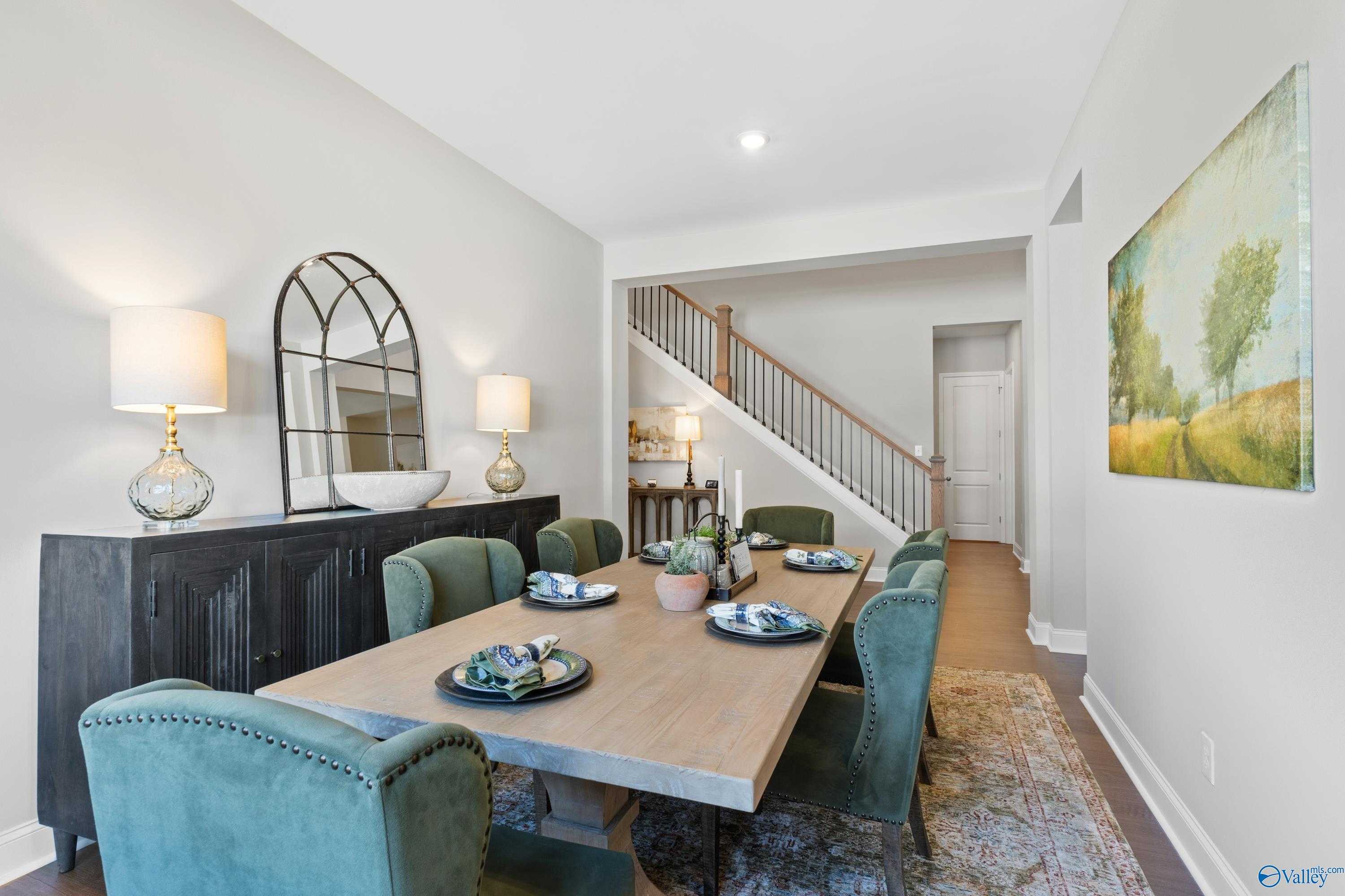 Elegant dining room with farmhouse table, emerald green velvet chairs, arched mirror, and staircase in Evermore Homes The Haven, Owens Cross Roads, Alabama