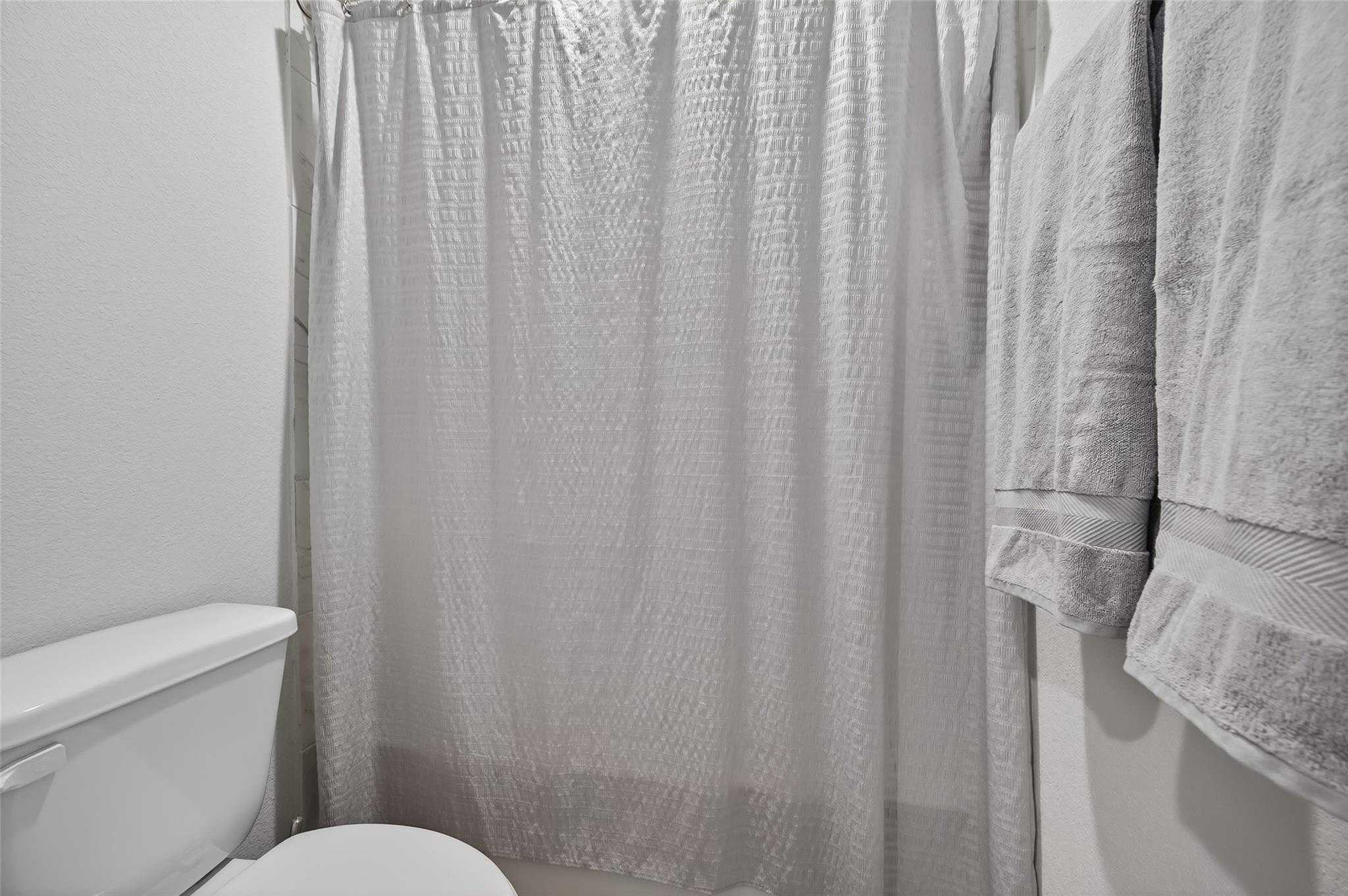 Modern bathroom with white textured shower curtain, toilet, and folded towels in Davidson Homes The Brazos E, Magnolia Texas
