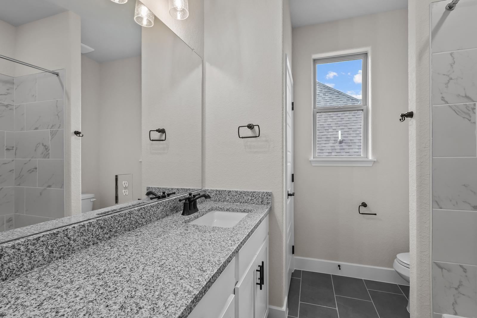 Modern bathroom in The Belmont featuring granite vanity, white cabinetry, and tiled shower in Castroville, Texas.