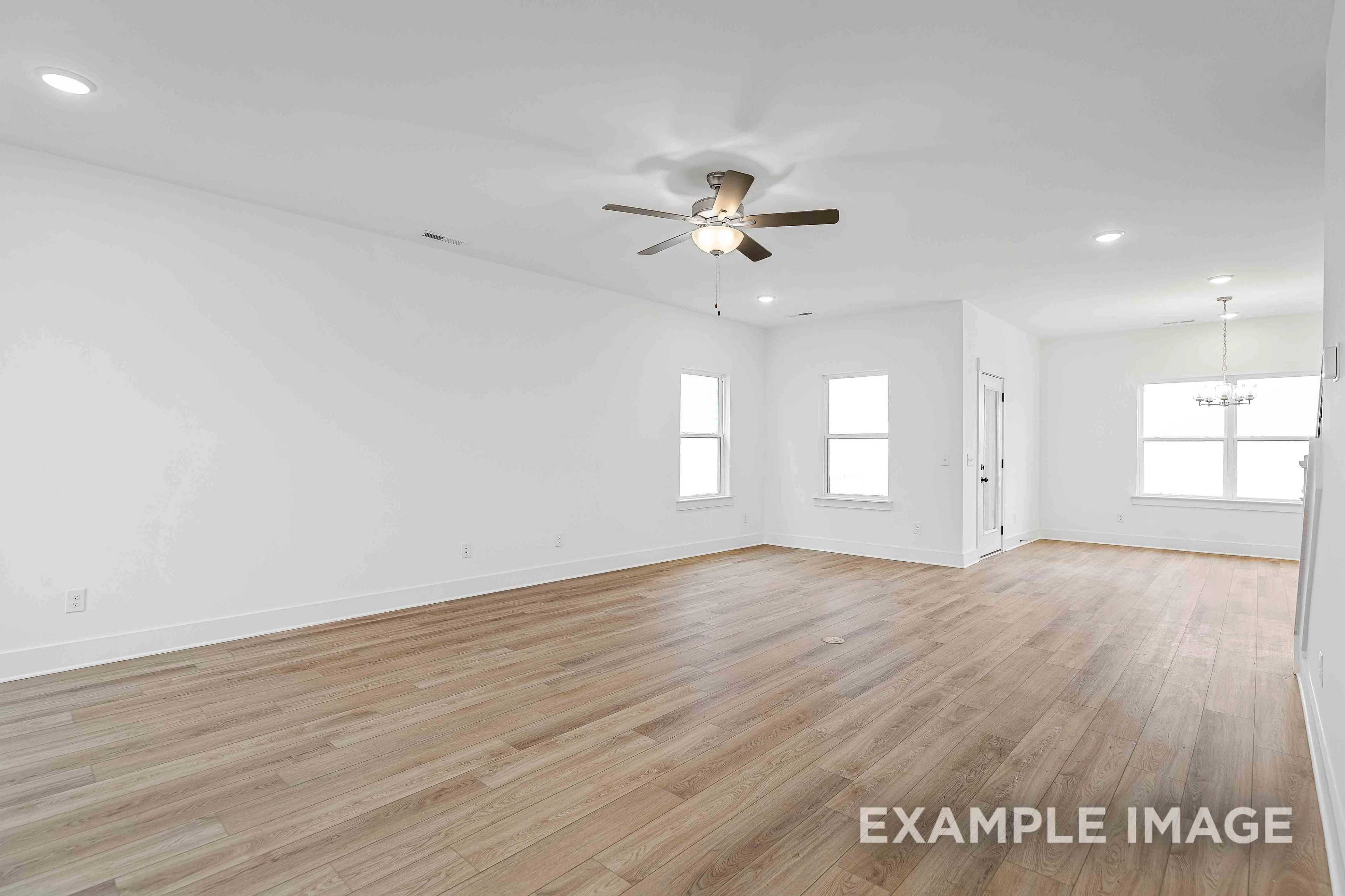 Spacious empty living room in The Charleston home plan with hardwood floors, white walls, ceiling fan, and large windows