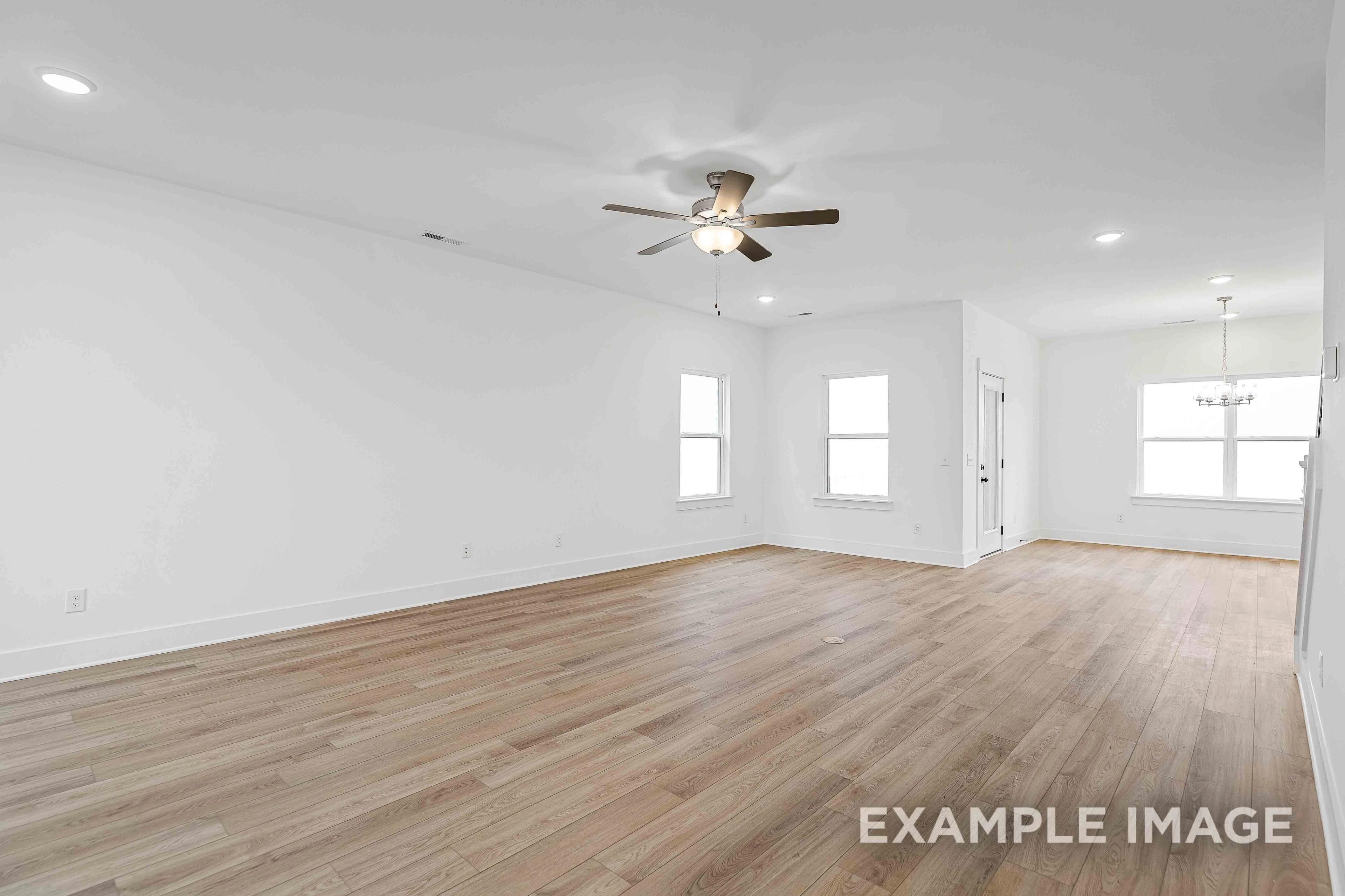Spacious empty living room in The Charleston home plan with hardwood floors, white walls, ceiling fan, and large windows
