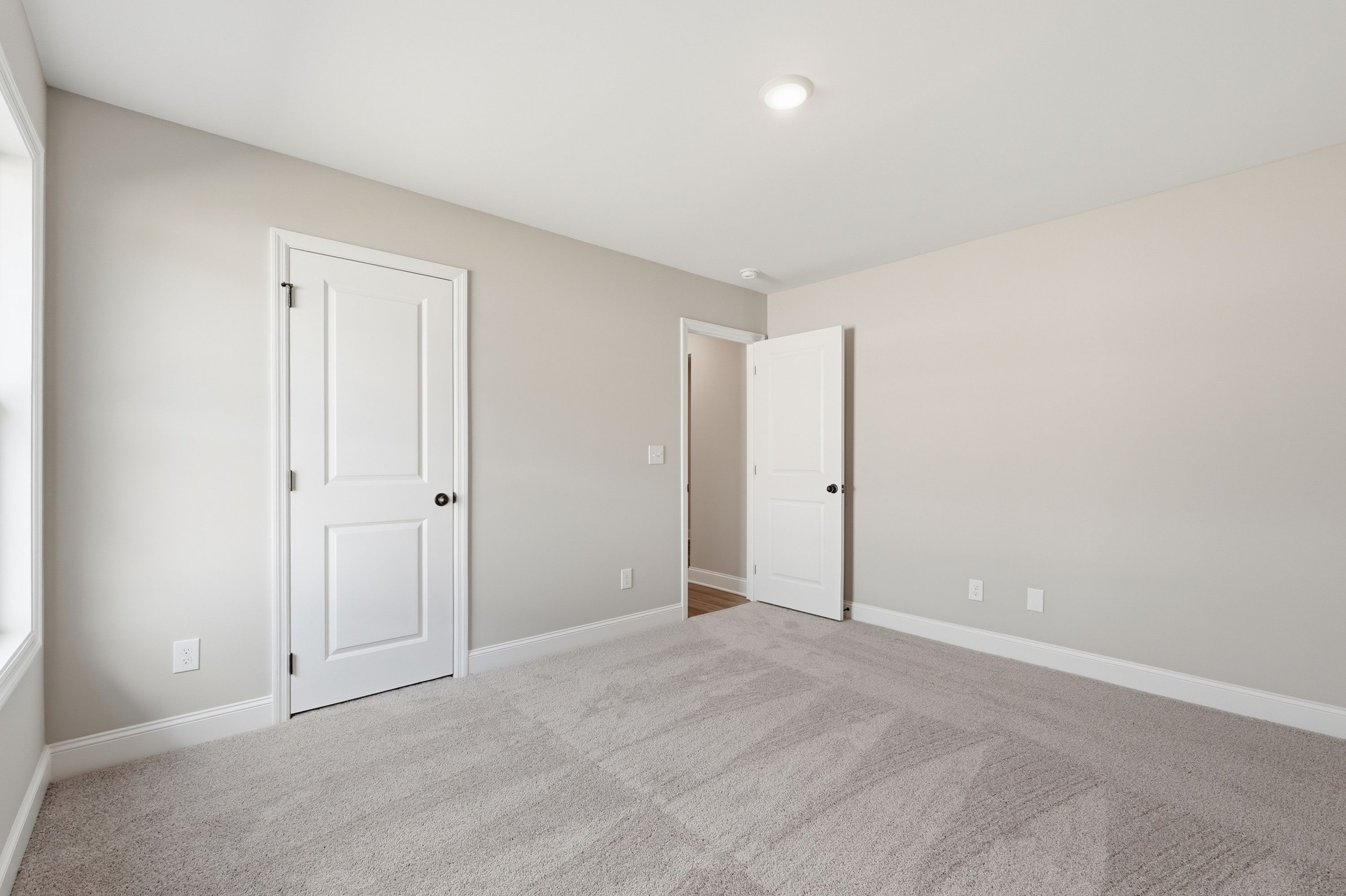 Spacious bedroom in The Daphne V with neutral beige walls, white doors, gray carpet, and large window in Meridianville home