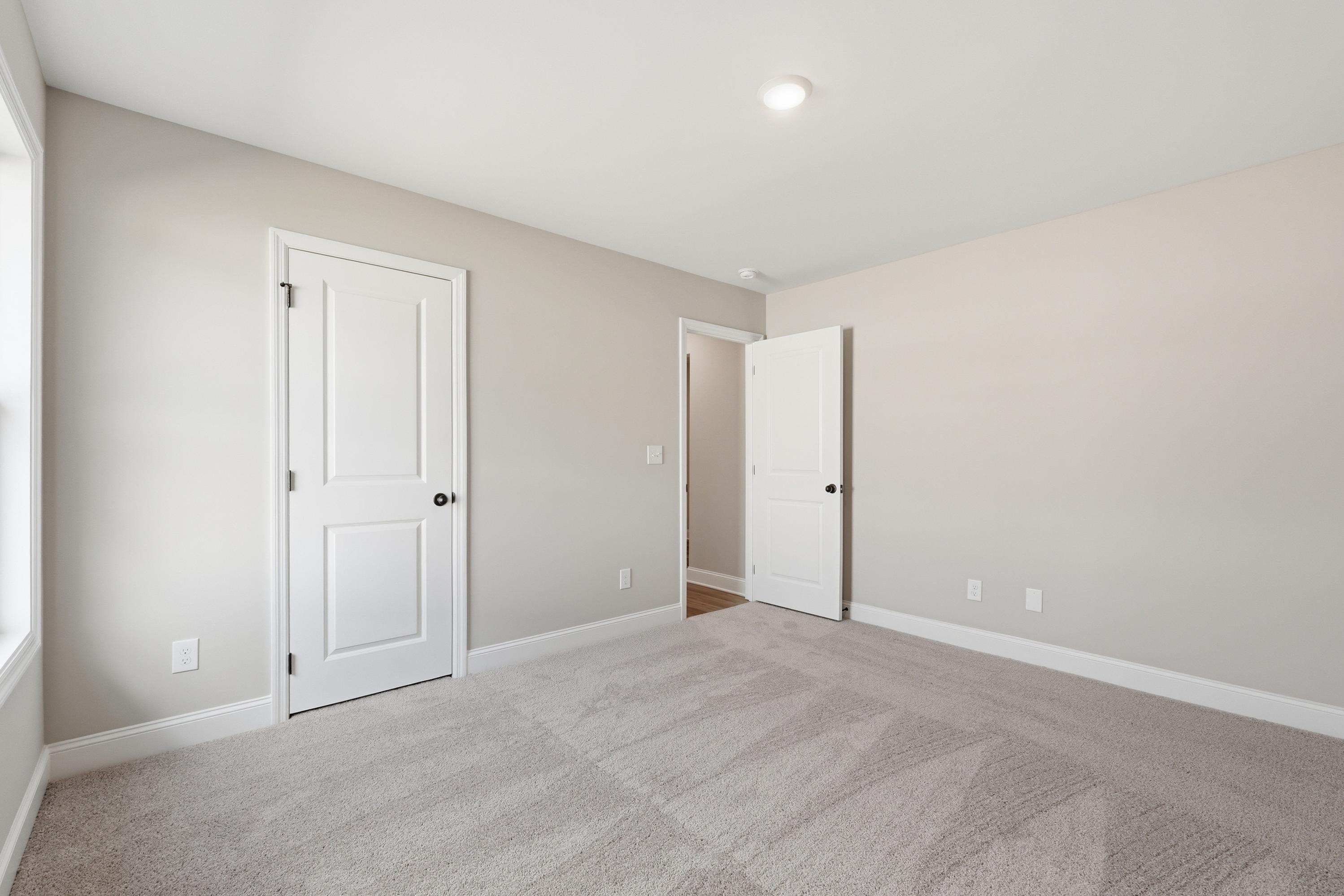 Spacious bedroom in The Daphne V with neutral beige walls, white doors, gray carpet, and large window in Meridianville home